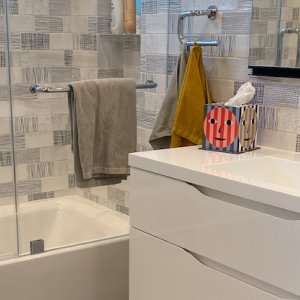 Modern bathroom with tiled walls, white vanity, colorful tissue box, and towels on rod near bathtub.