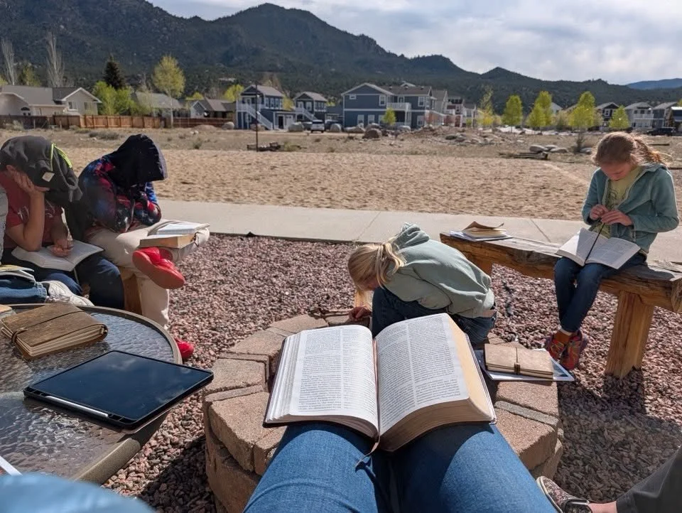 Group of children reading and studying outdoors in a mountainous area with modern houses in the background.