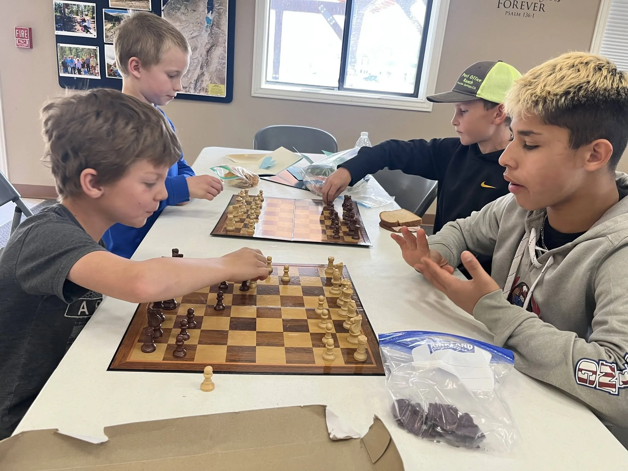 Four boys are playing chess at a table in a room with windows and pictures on the wall. The table has a chessboard and some snacks. One boy is adjusting the chess pieces, while the others are watching.