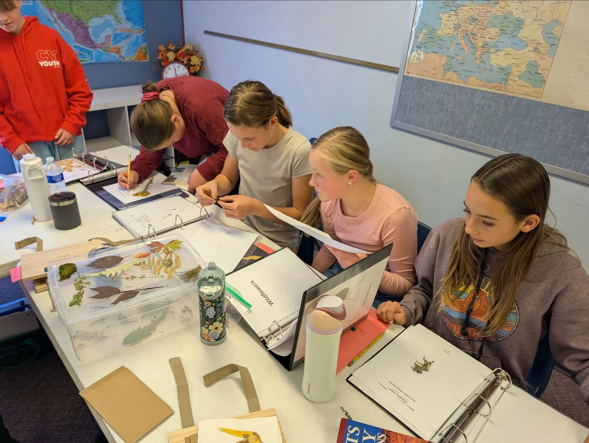 Group of children seated at a table with maps of the world on the walls, working on educational activities involving dried leaves and natural materials.
