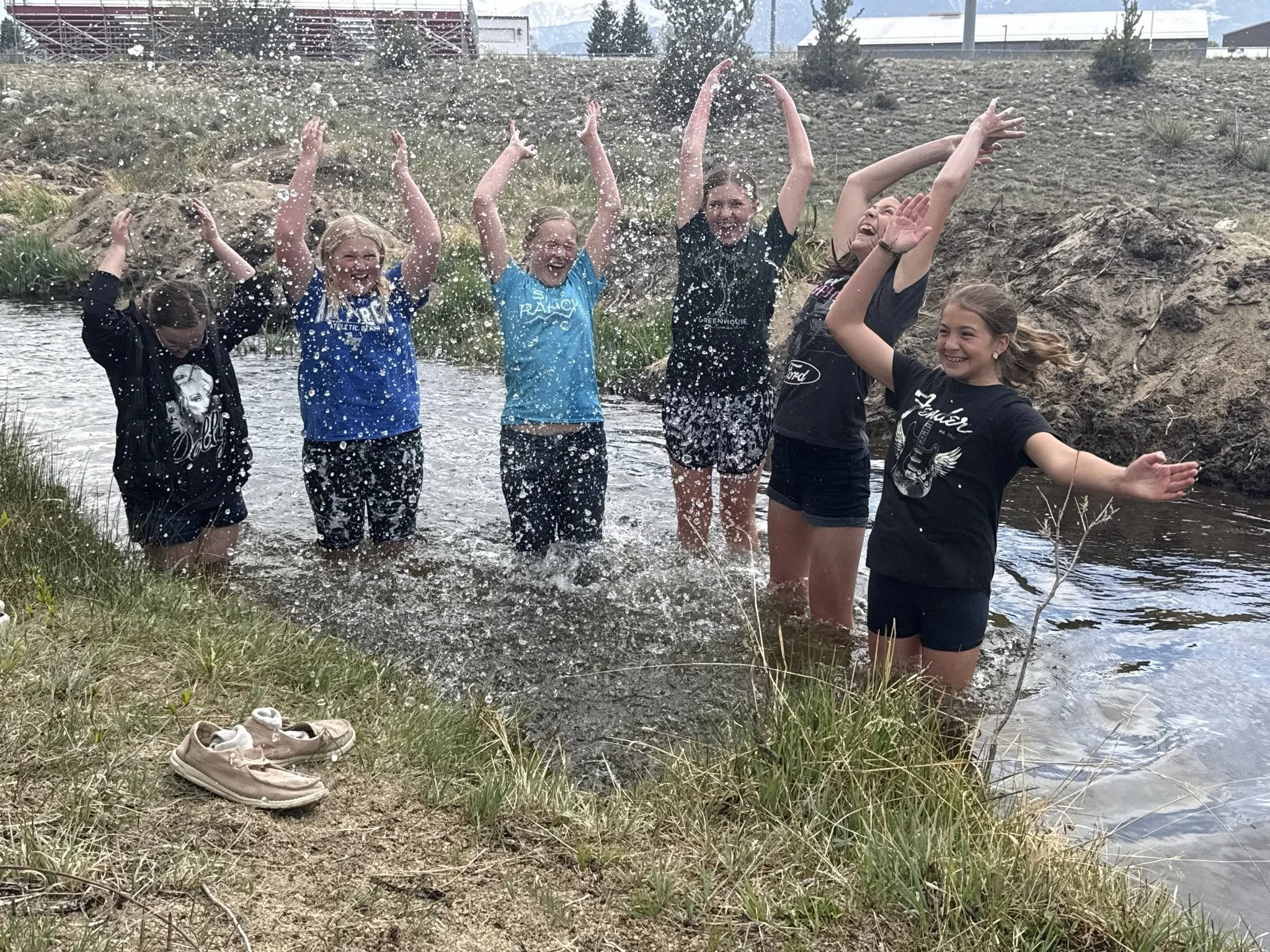 Six young girls standing in a shallow body of water, celebrating and splashing water in an outdoor setting with rocky terrain and some buildings in the background.