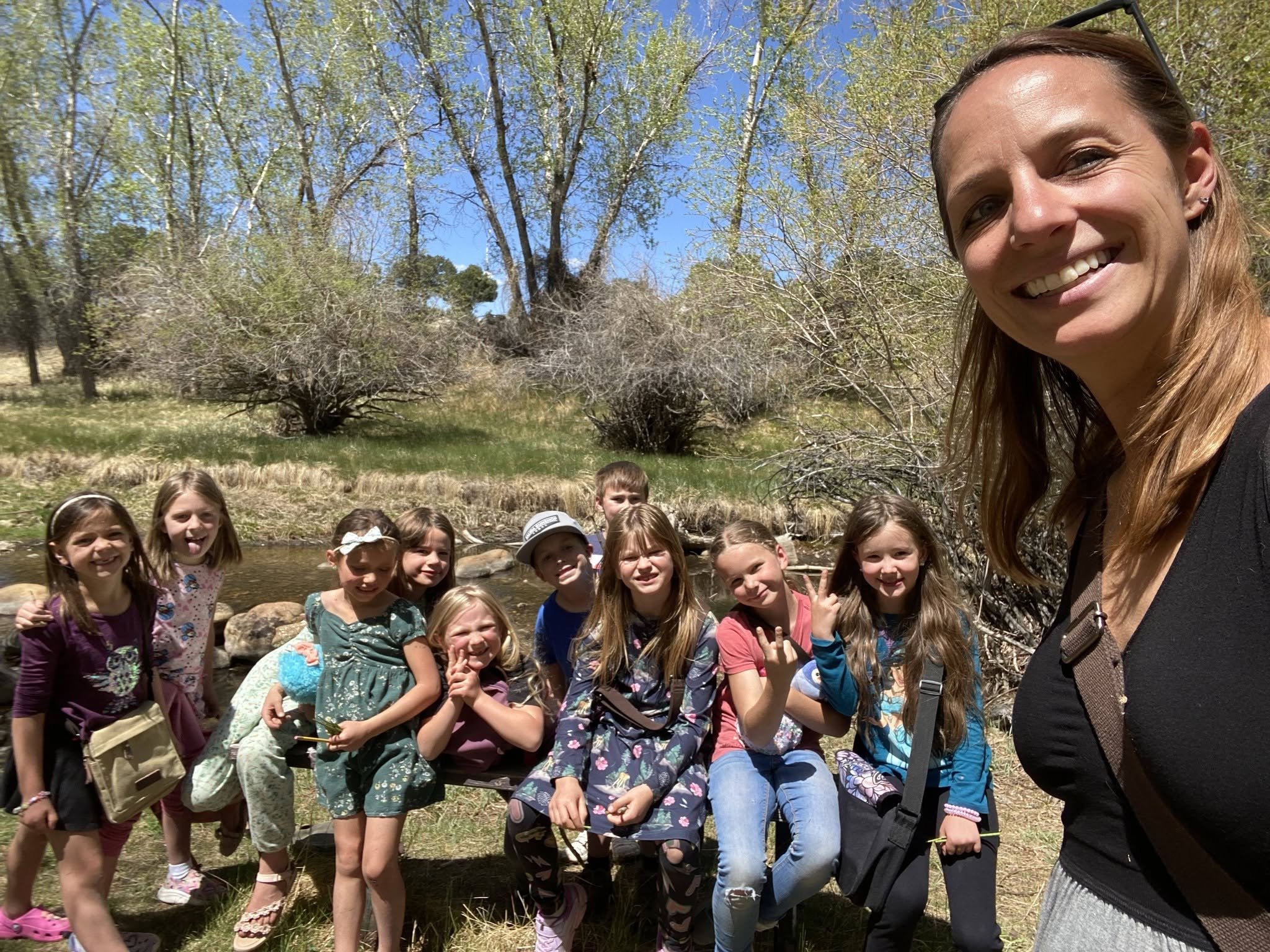 A woman takes a selfie with a group of children outdoors near a river, trees, and a blue sky.