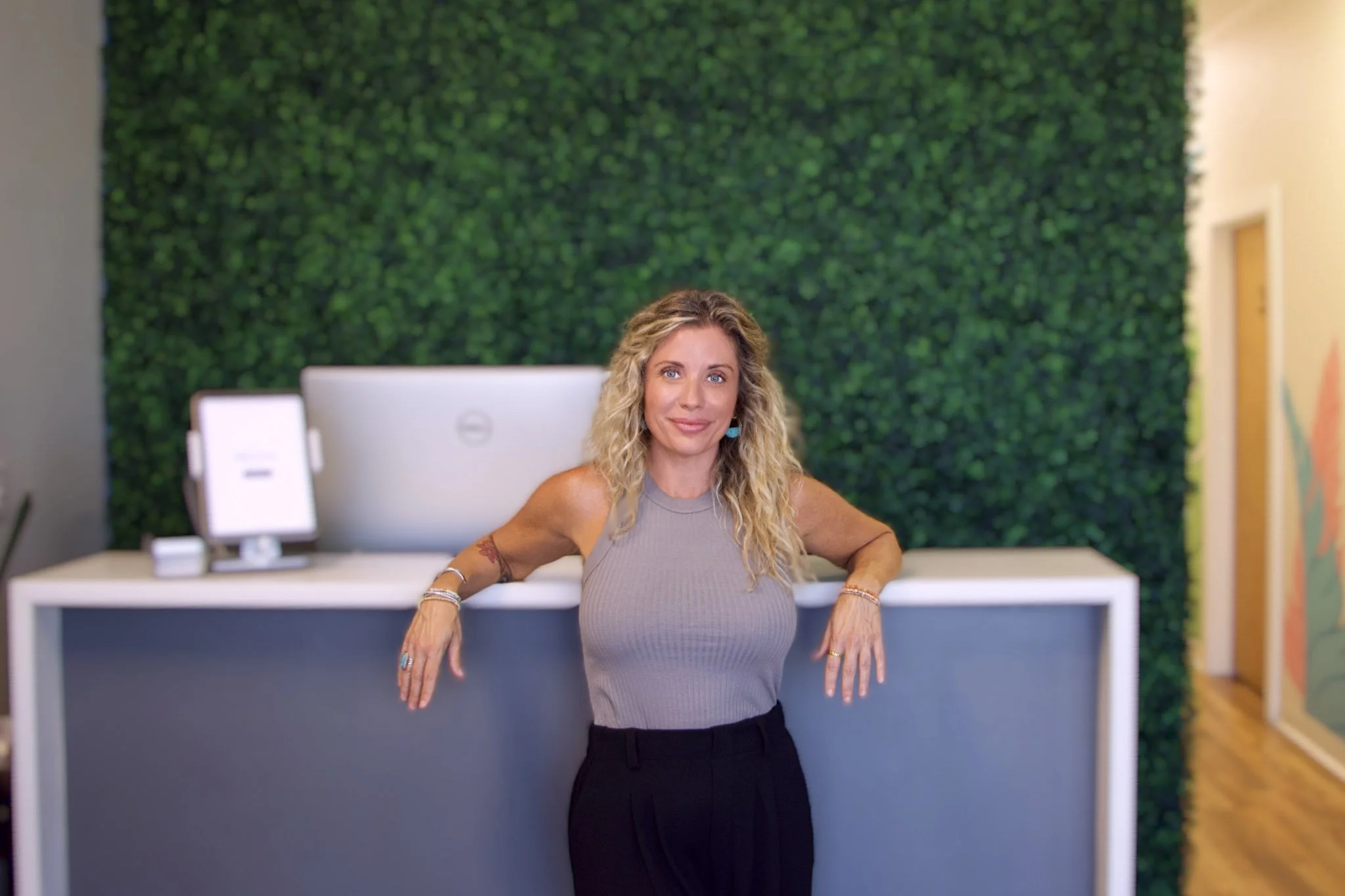A woman with blonde hair sitting on a white chair in front of a leafy green mural, wearing a dark tank top and ripped jeans.