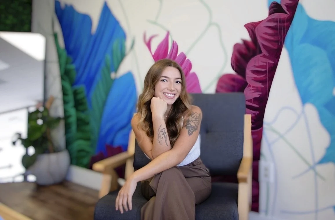 A woman sitting on a modern white chair with wooden legs, smiling, with her arms resting on the back of the chair. She has shoulder-length brown hair, tattoos on her left arm, and is wearing a black t-shirt and light blue jeans. She is barefoot with painted toenails. Behind her is a decorative wall with large green tropical leaves, and to her left is a tall potted snake plant in a beige patterned pot. The floor is wooden, and there is a white electrical outlet and a switch on the wall behind her.