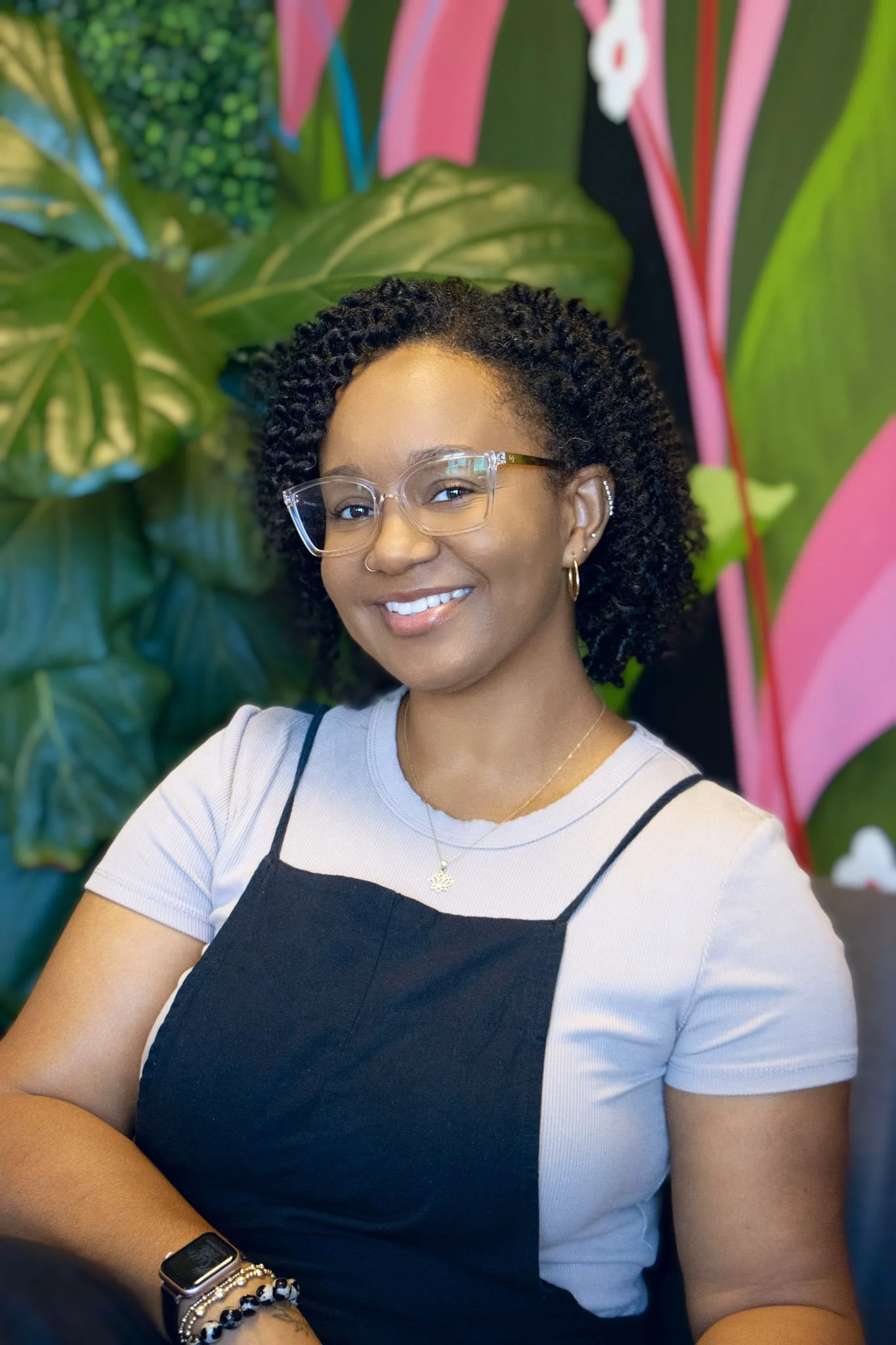 A woman with glasses and braided hair sitting in front of a tropical leaf-patterned wallpaper, wearing a white shirt and light blue ripped jeans, smiling at the camera.
