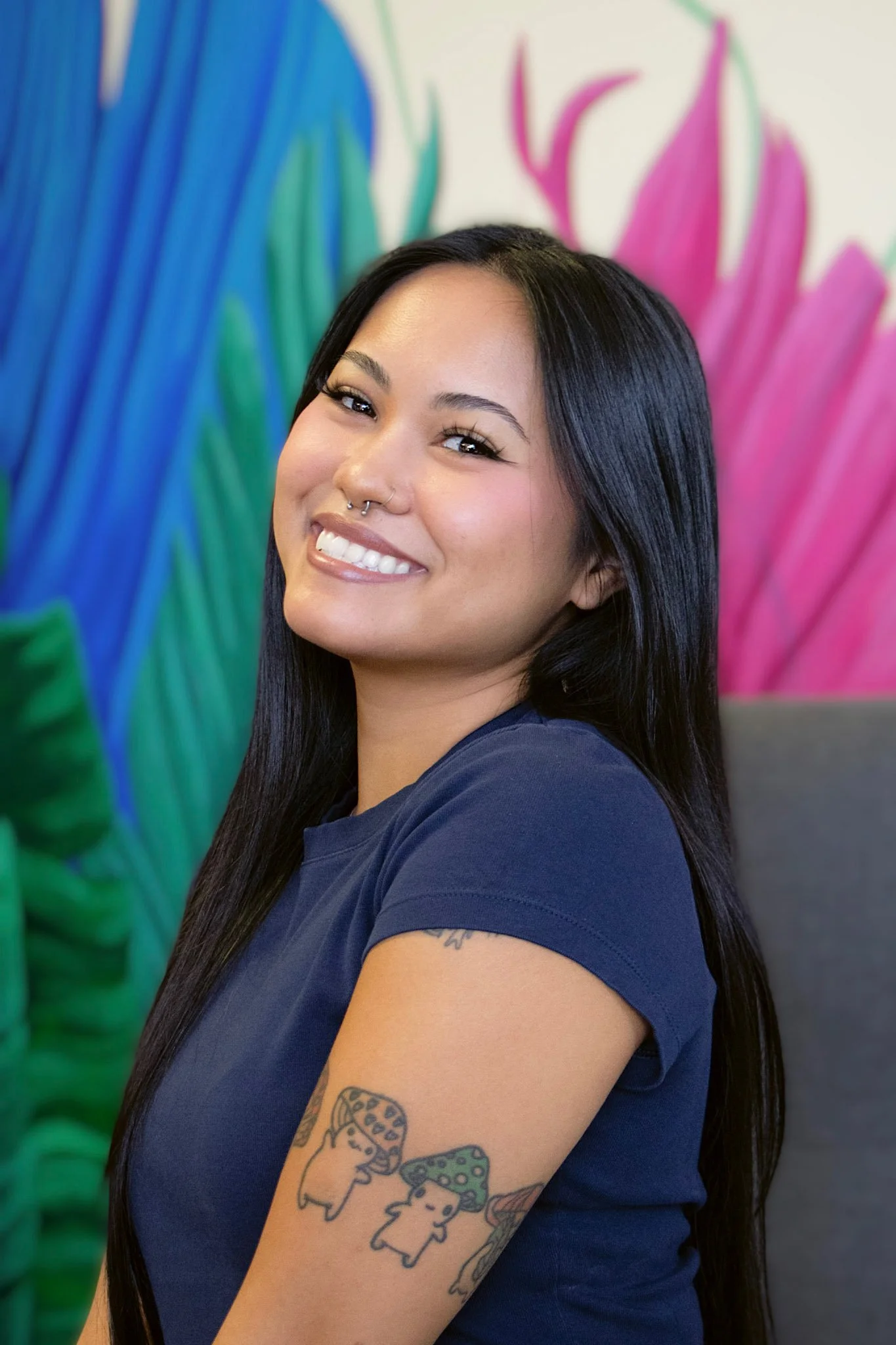 A young woman with long dark hair and tattoos on her arm, smiling and sitting on a white chair in front of a leafy green wallpaper and a large circular mirror, with potted plants nearby.