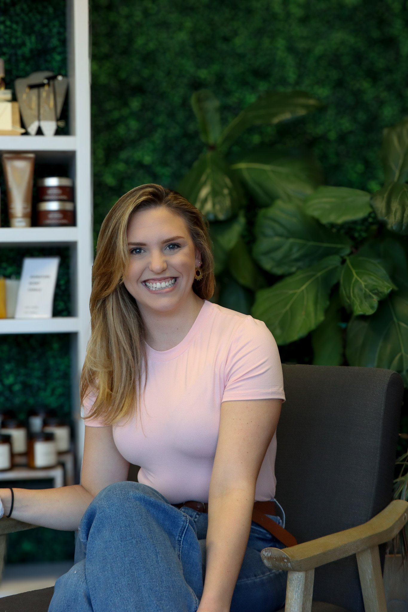 A woman with long hair, smiling, sitting in front of a wall decorated with green tropical leaves, wearing a pink top and jeans.