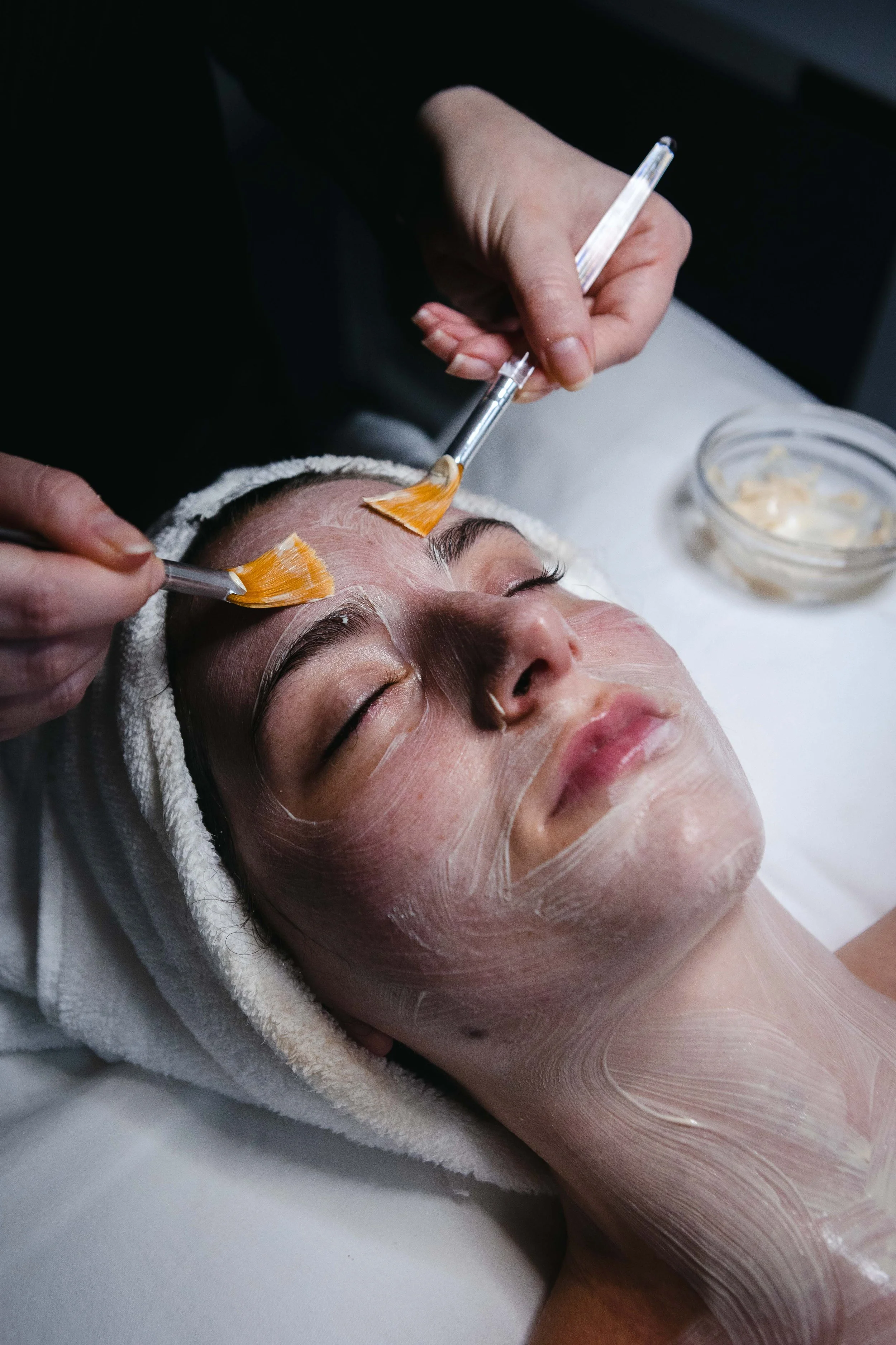 A woman receiving a facial treatment at a spa, lying on a bed with her eyes closed, while beauticians apply a facial mask with brushes.