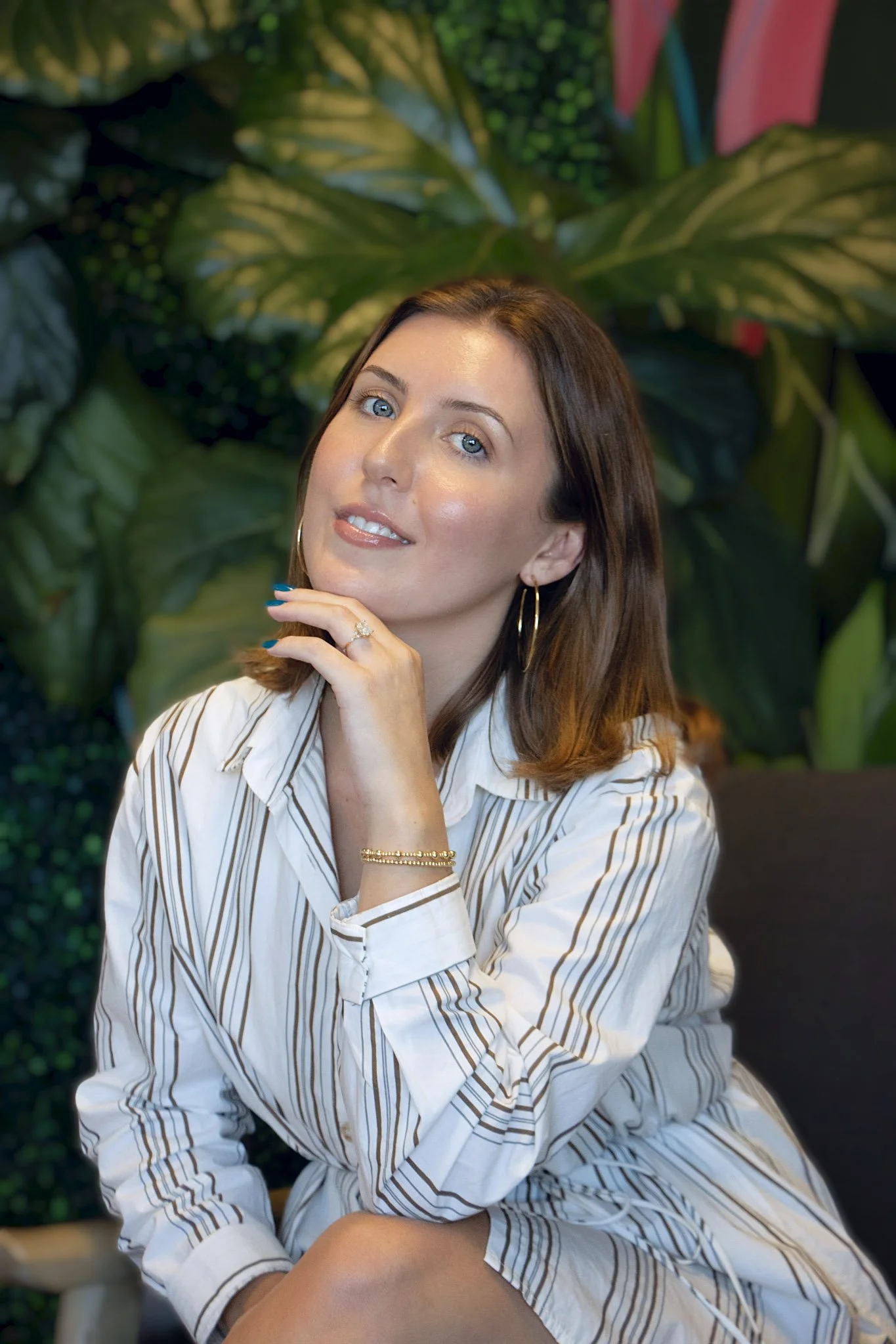 A young woman with long brown hair smiling, sitting on a white chair with one arm resting on her knee, in front of a green leafy tropical wallpaper, wearing a white t-shirt, ripped jeans, and sneakers.