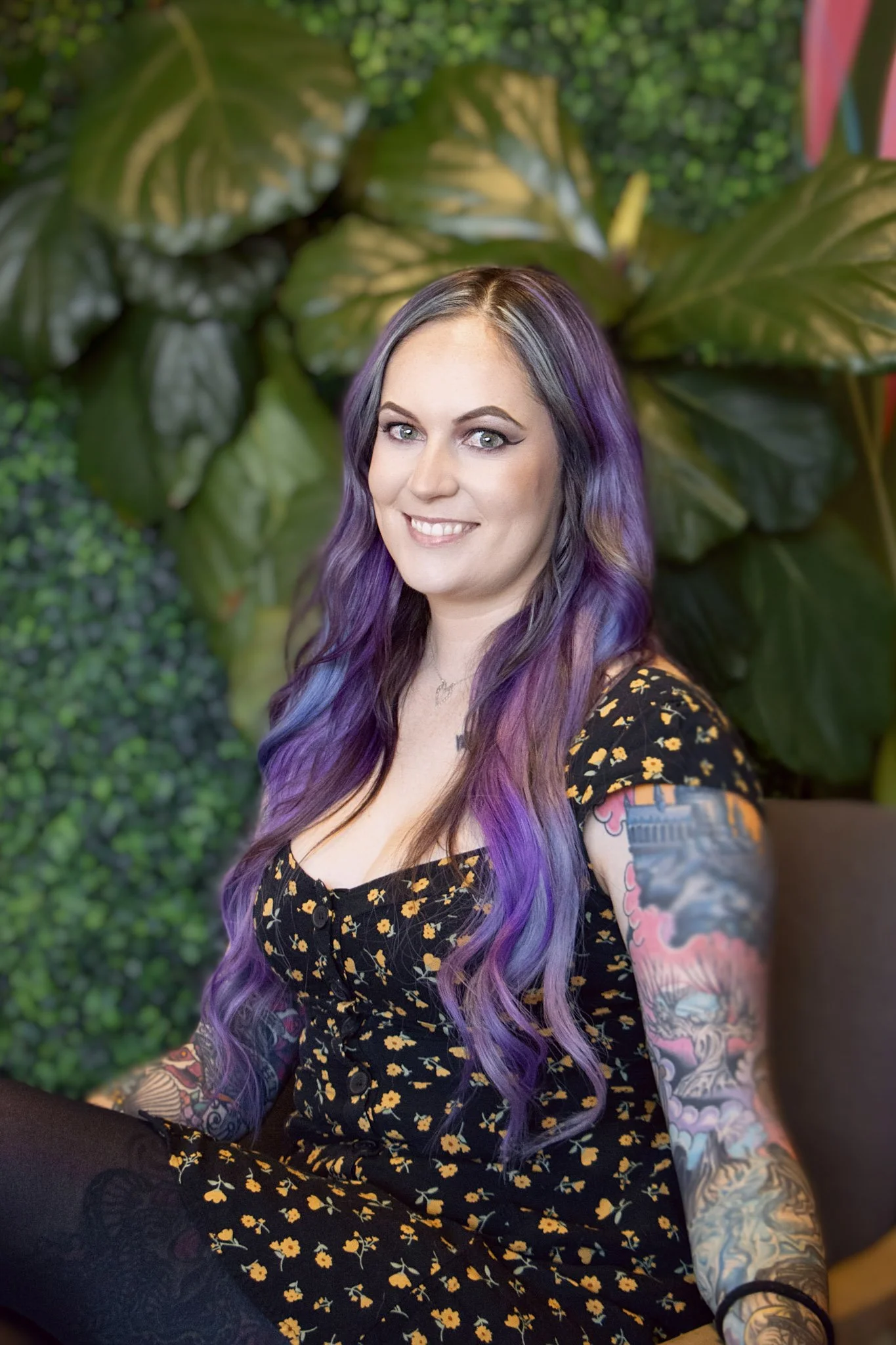 A smiling woman with black and purple hair, tattoos on her arms, wearing a white sleeveless top and olive green pants, sitting on a white chair in front of a leafy green plant wallpaper.