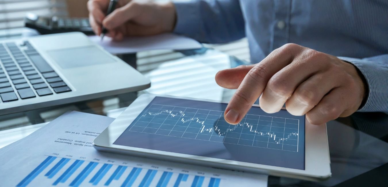 Person using a tablet with a financial chart, surrounded by a laptop, papers, and a notepad on a glass conference table.