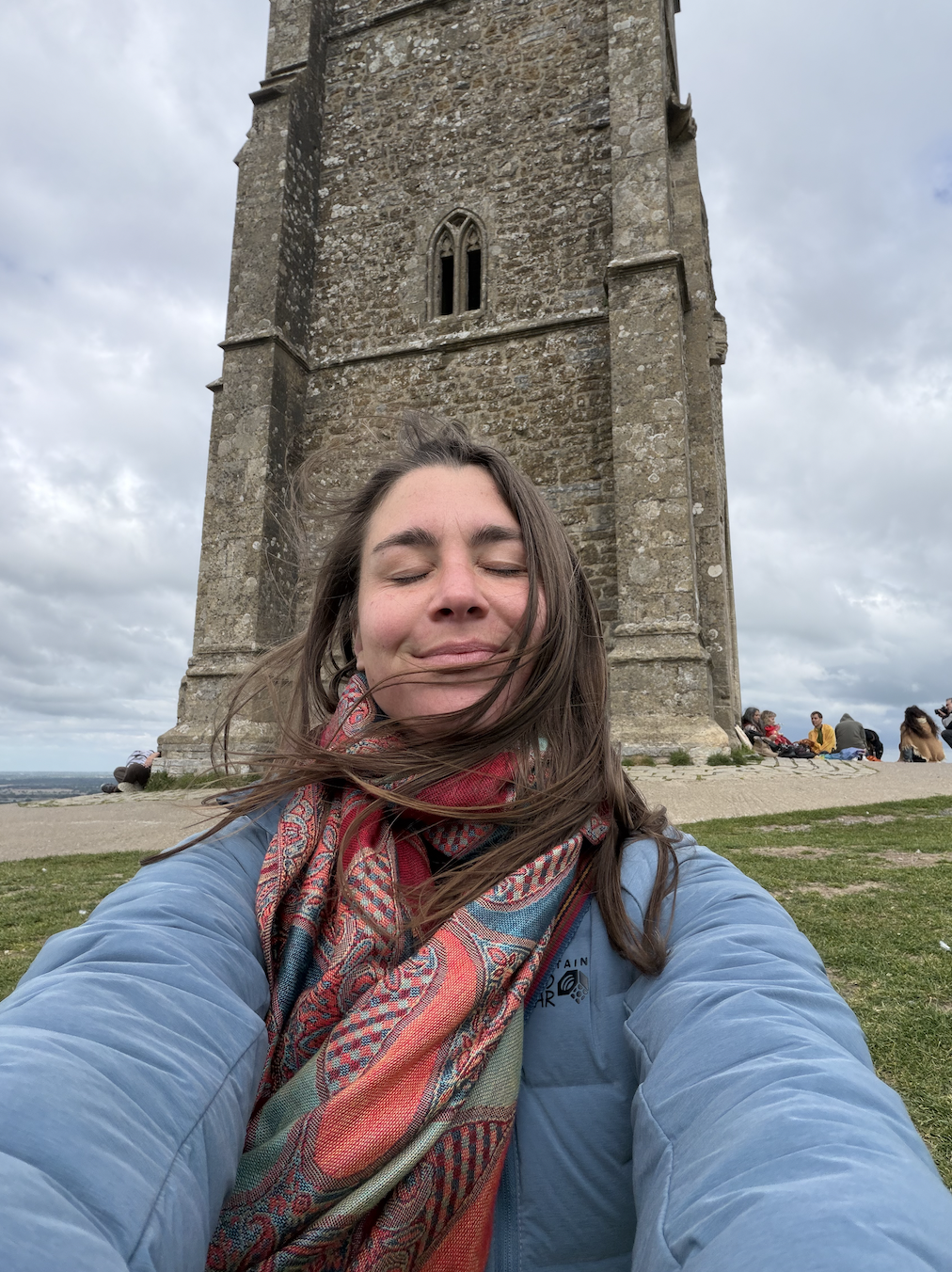 A woman taking a selfie in front of a tall, stone clock tower with a cloudy sky. She has closed eyes, wind-blown hair, and is smiling, wearing a colorful scarf and a light blue jacket.