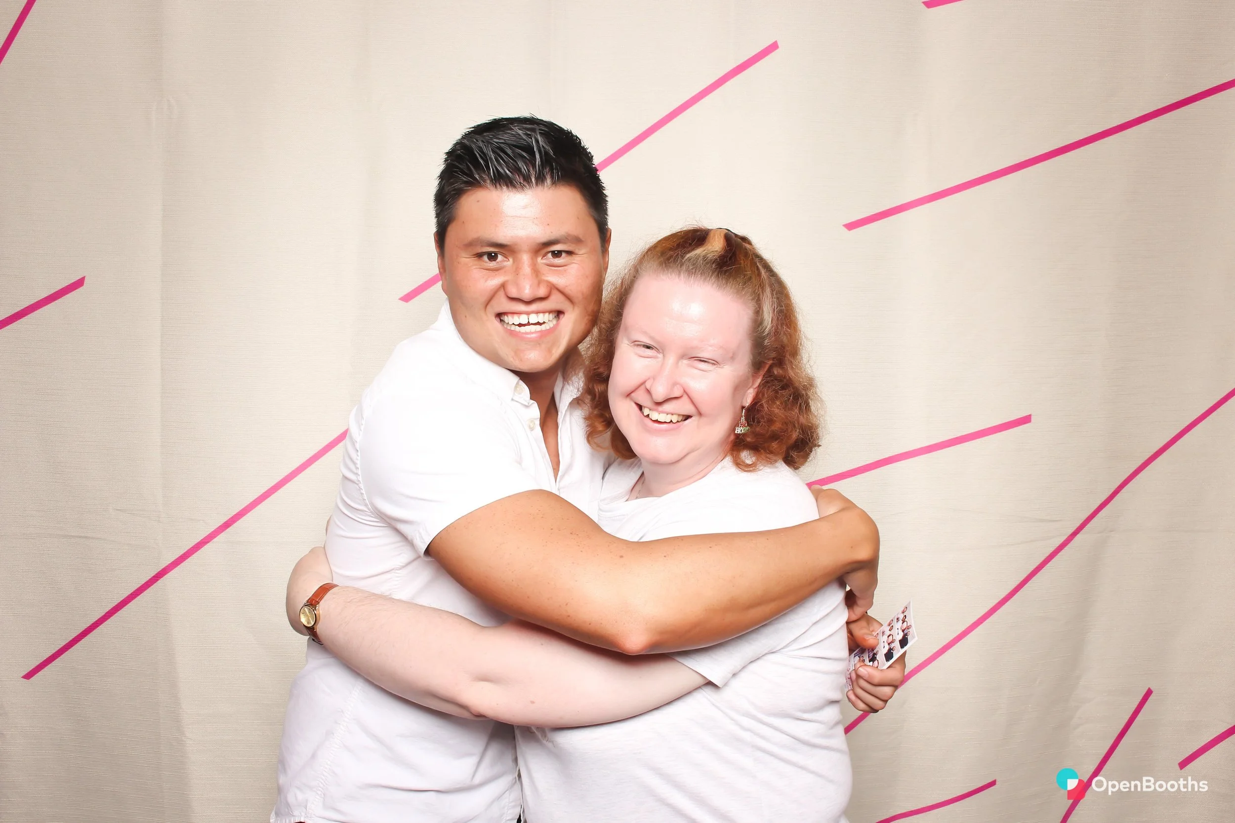 Two people pose in an Open Booths photo booth with a cream backdrop featuring hot pink diagonal lines