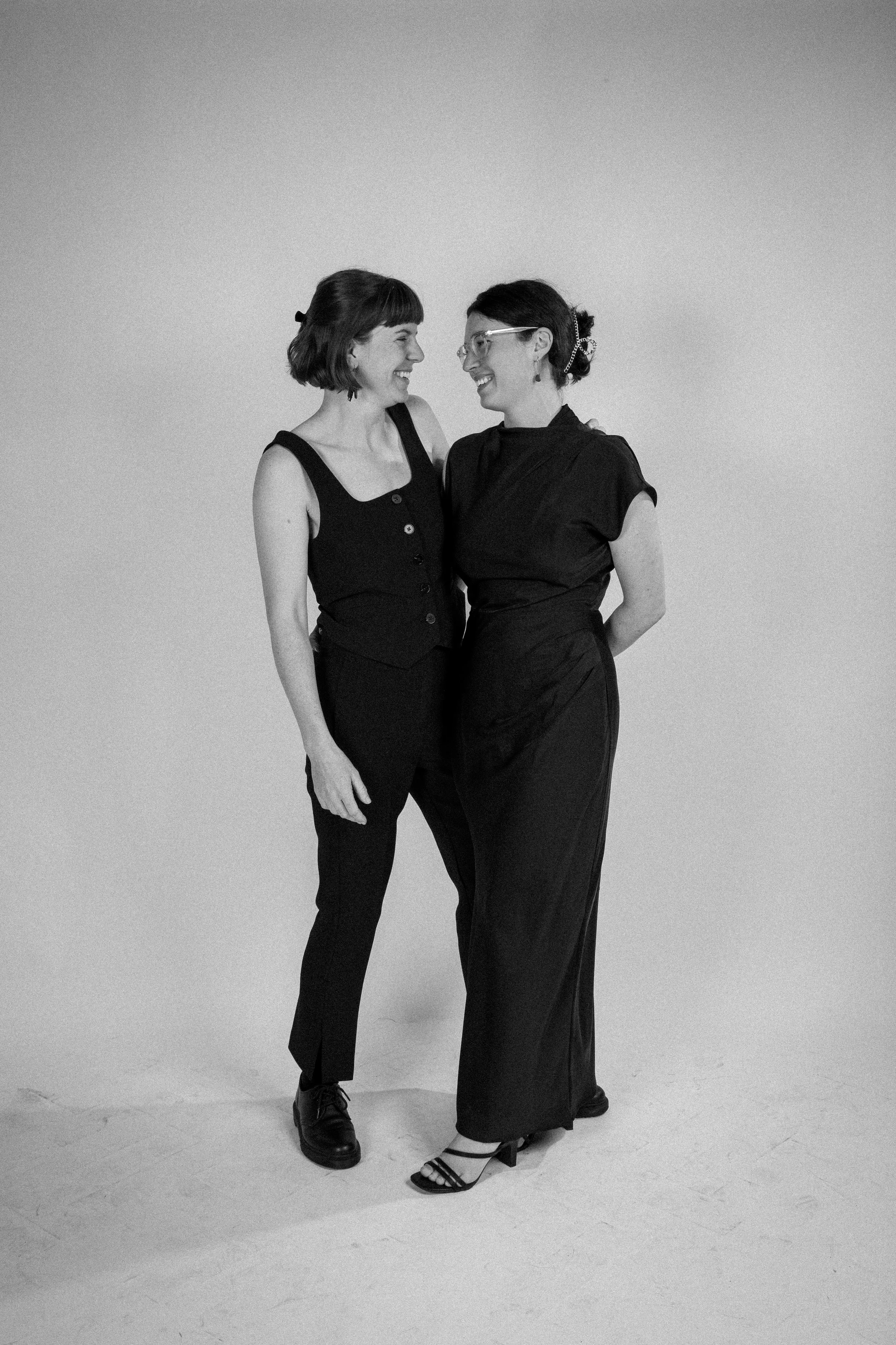 A studio portrait photo of two wedding guests turning to face one another in front of a white paper infinity wall.