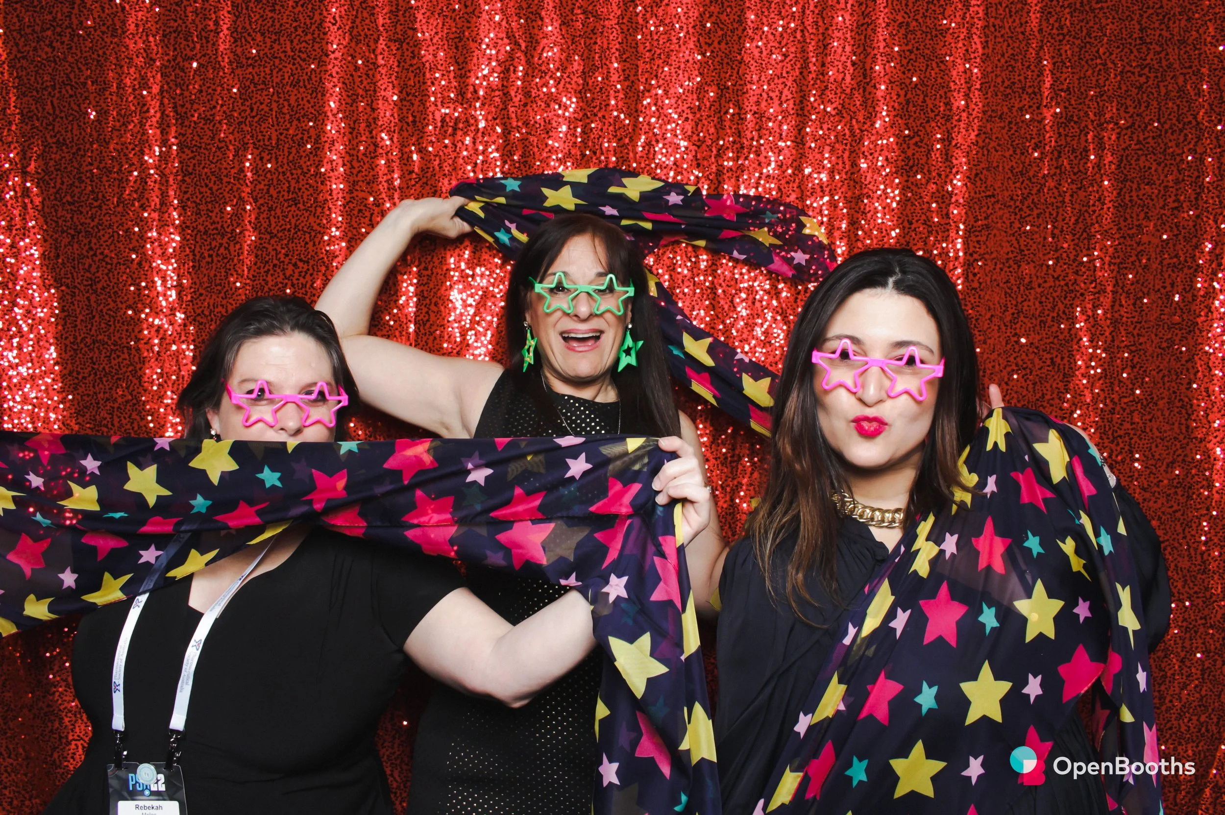 Three women play with star printed fabric in an Open Booths photo booth featuring a ruby red sequin backdrop