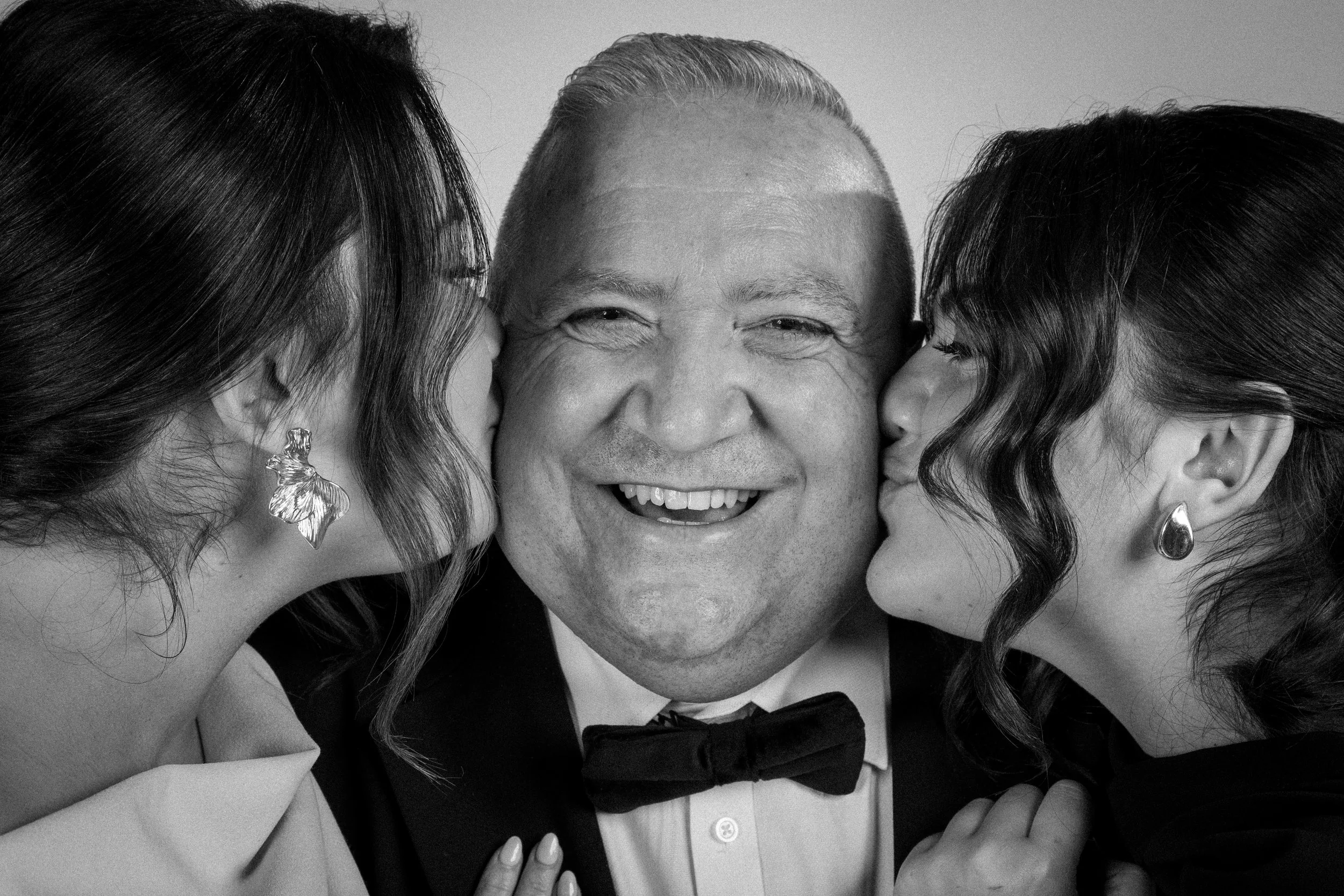 Man smiling broadly while receiving kisses on the cheeks in a joyful family wedding portrait.