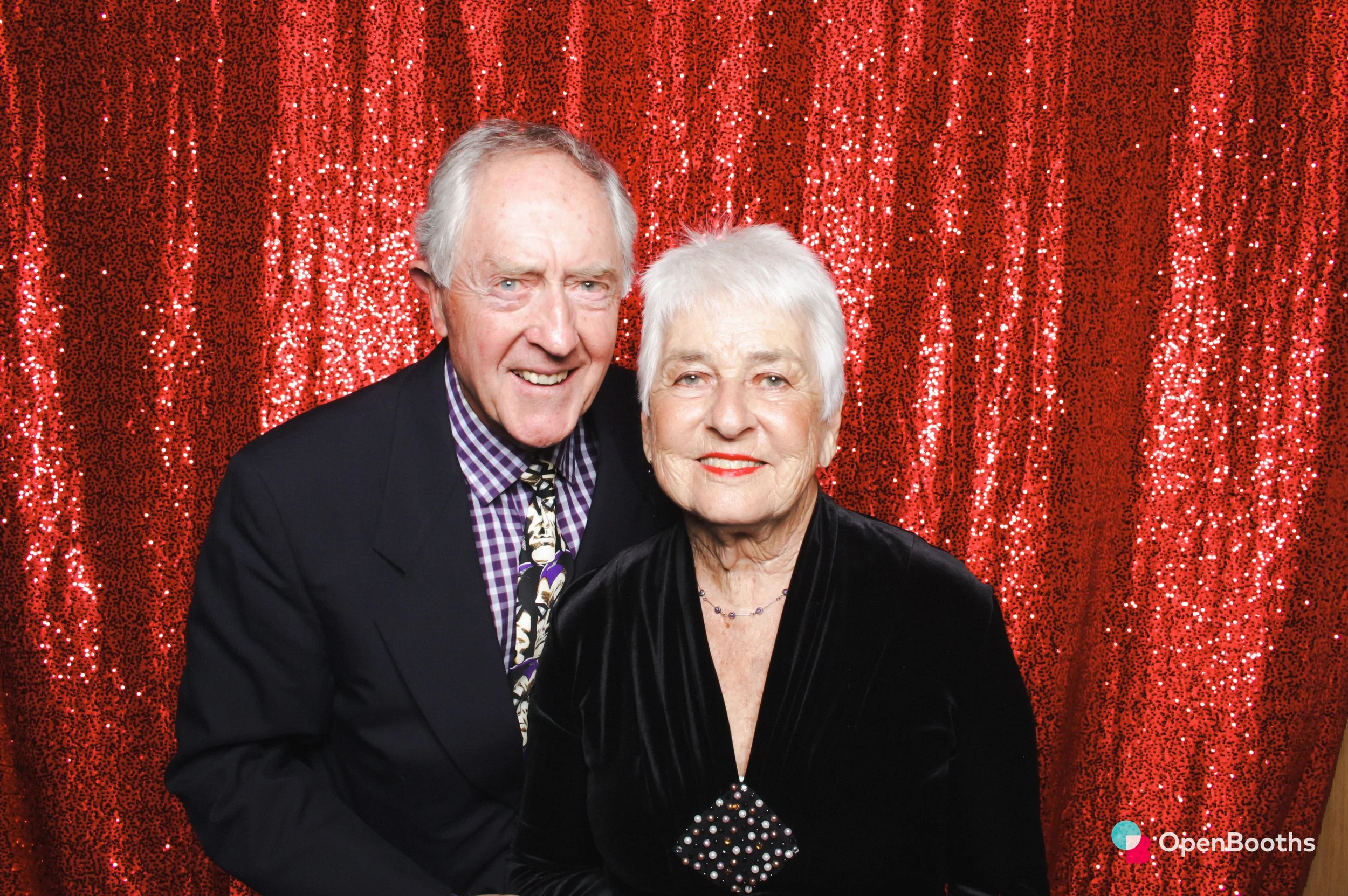 A couple pose for a photo in an Open Booths photo booth with a sparkly red sequin backdrop