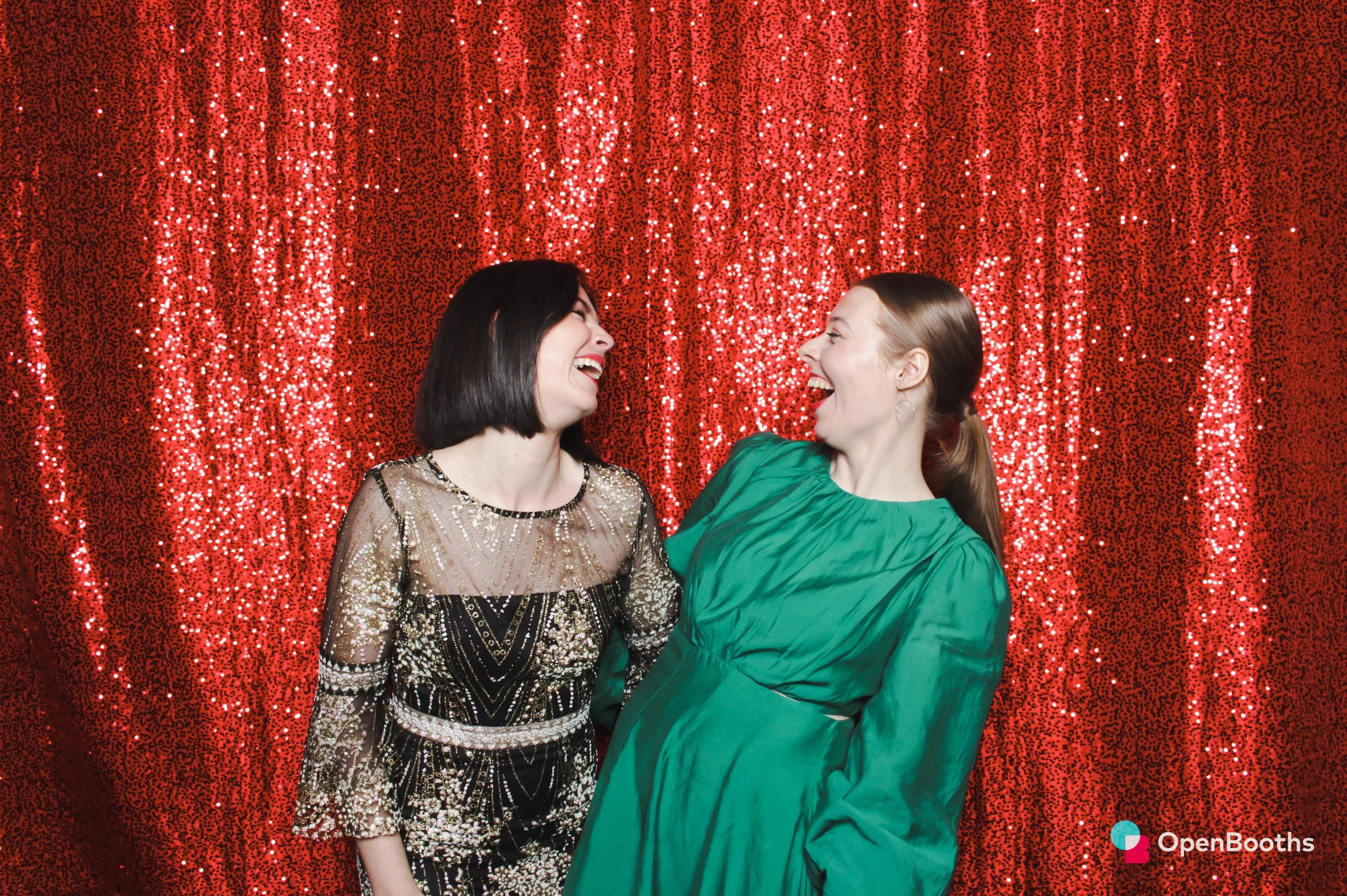 Two women smile at each other in an Open Booths photo booth featuring a red sequin backdrop