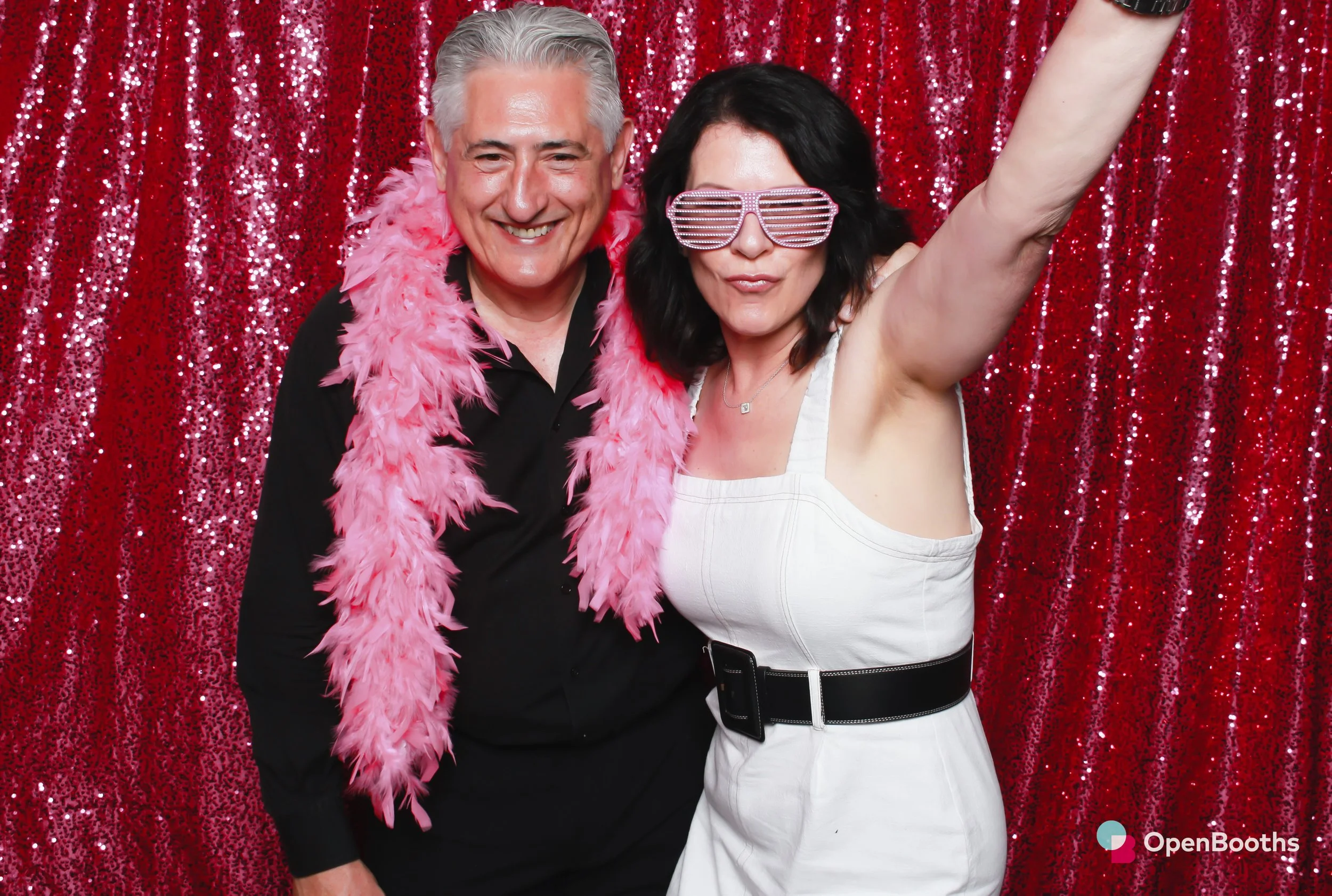 Open Booths photo booth image of a group of two people posing in front of a glittery pink sequin backdrop wearing shutter shades and a feather boa
