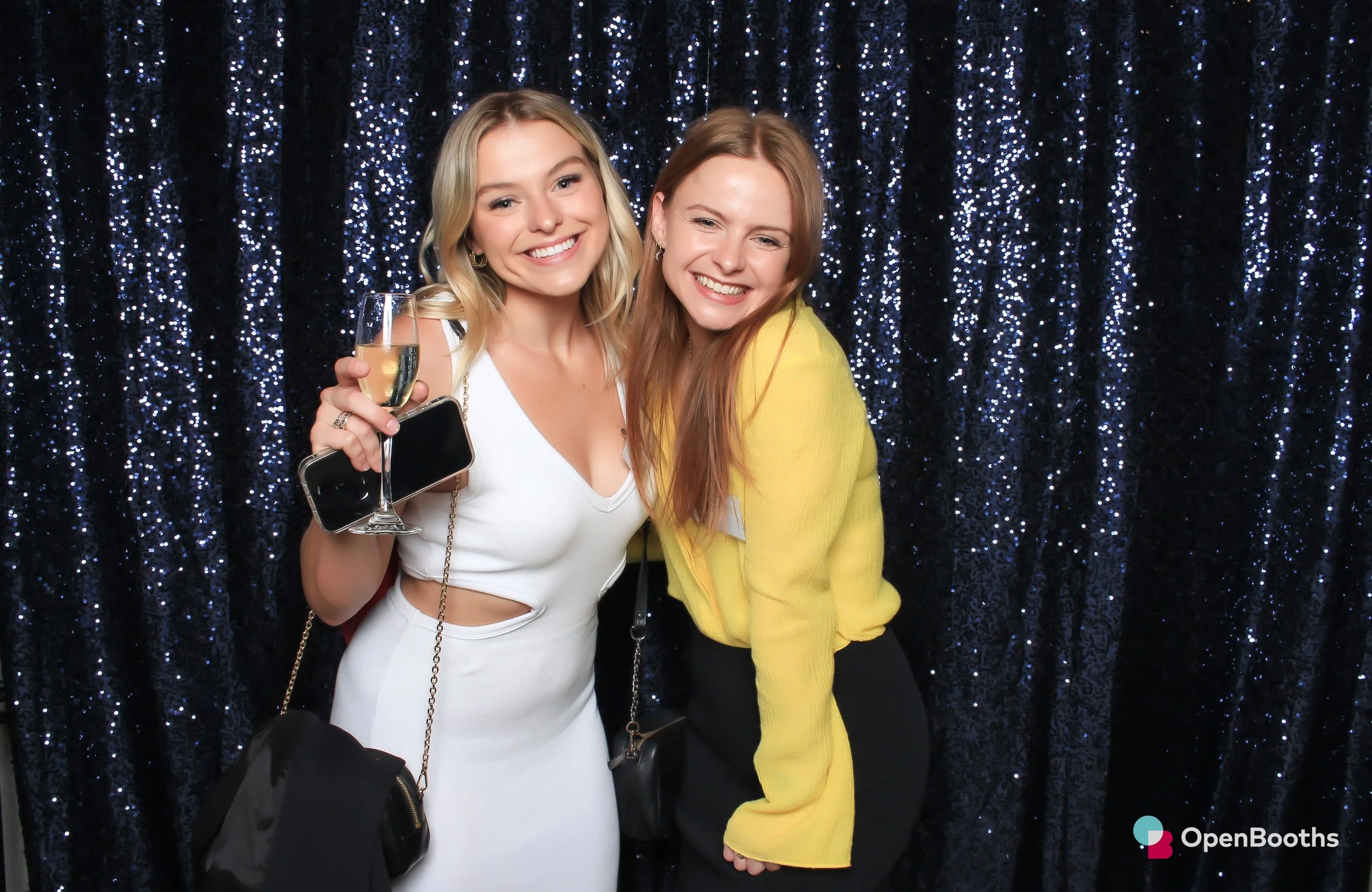 Two women pose in front of a deep navy blue sequin backdrop in an Open Booths photo booth