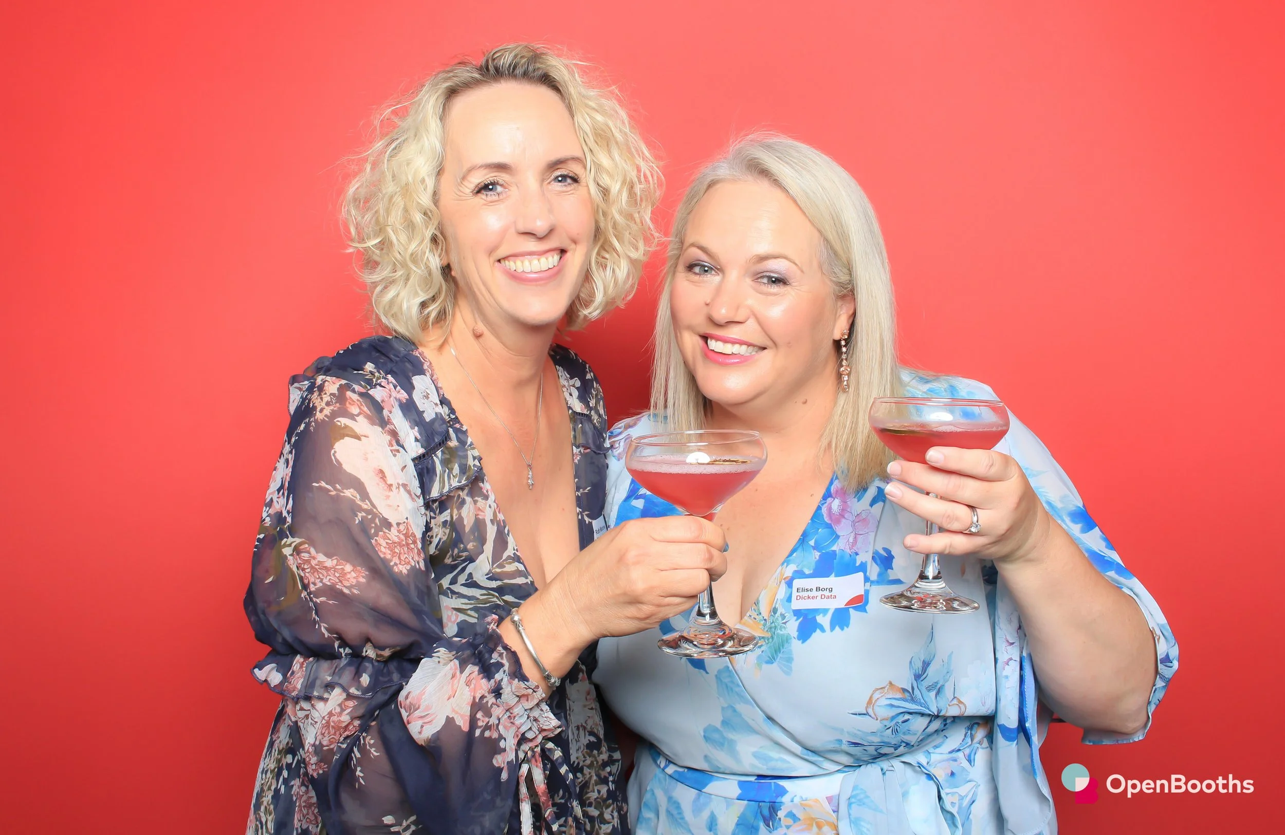 Two women stand in front of an Open Booths Racy Red backdrop and hold up red drinks