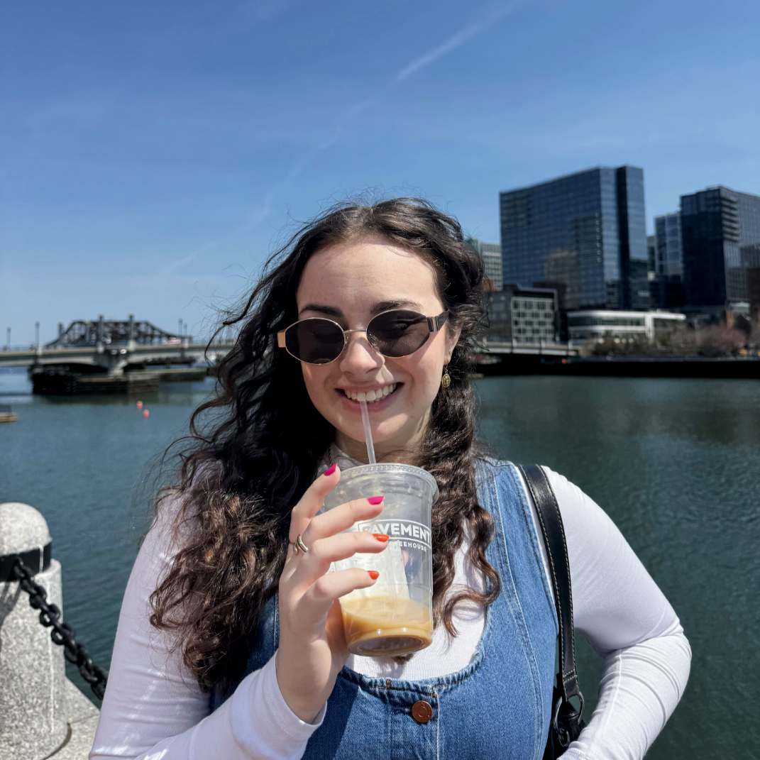 Young woman with dark curly hair wearing sunglasses, white shirt, and denim overalls, holding an iced coffee, smiling near a body of water with a cityscape of modern buildings and a bridge in the background on a sunny day.