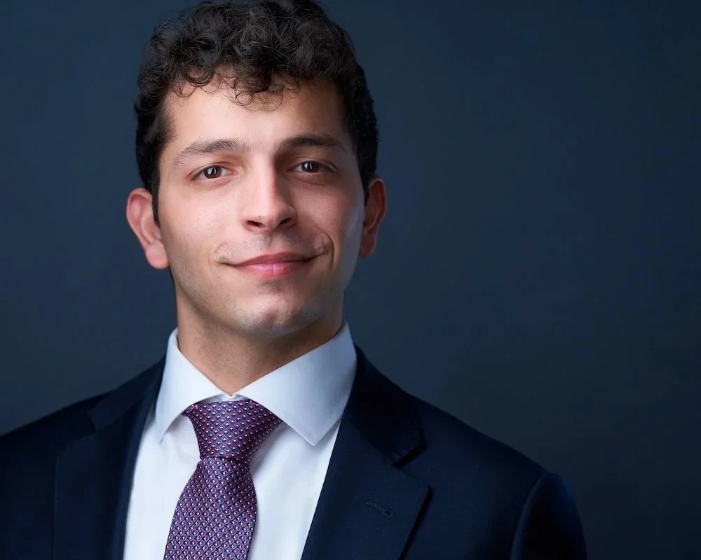 A young man in a dark suit, white shirt, and patterned tie, smiling slightly against a dark background.