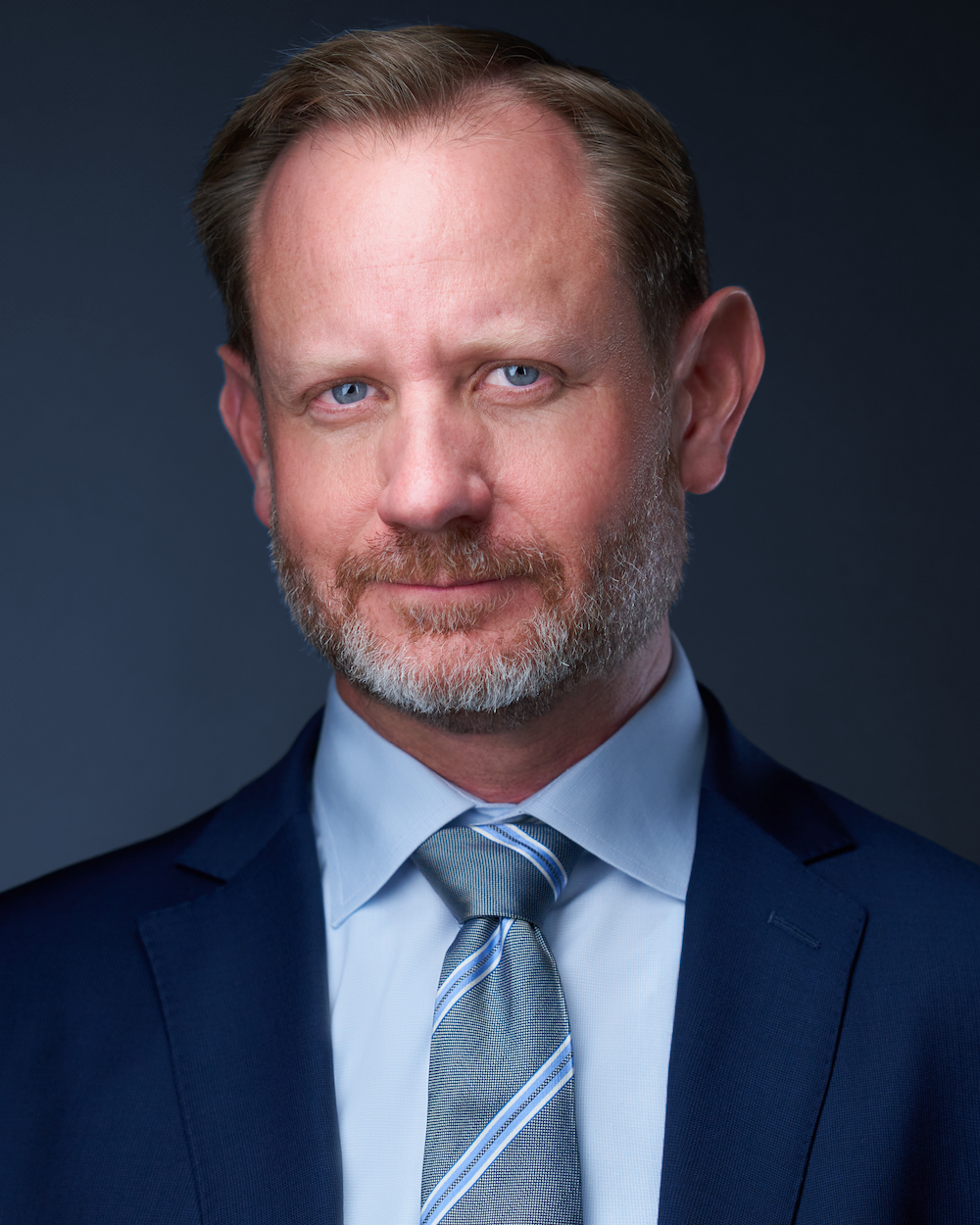 Close-up portrait of a middle-aged man with blue eyes, light brown hair, and a beard, dressed in a dark blue suit, white shirt, and a striped tie, against a dark background.