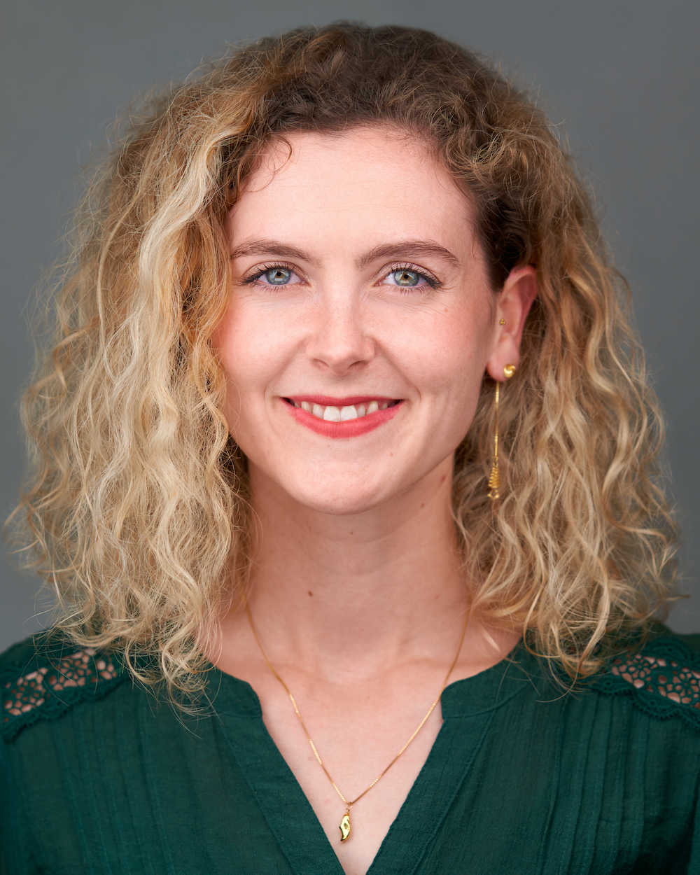 A woman with curly blonde hair, fair complexion, and blue eyes smiling at the camera, wearing earrings and a green blouse with lace detailing on the shoulders.