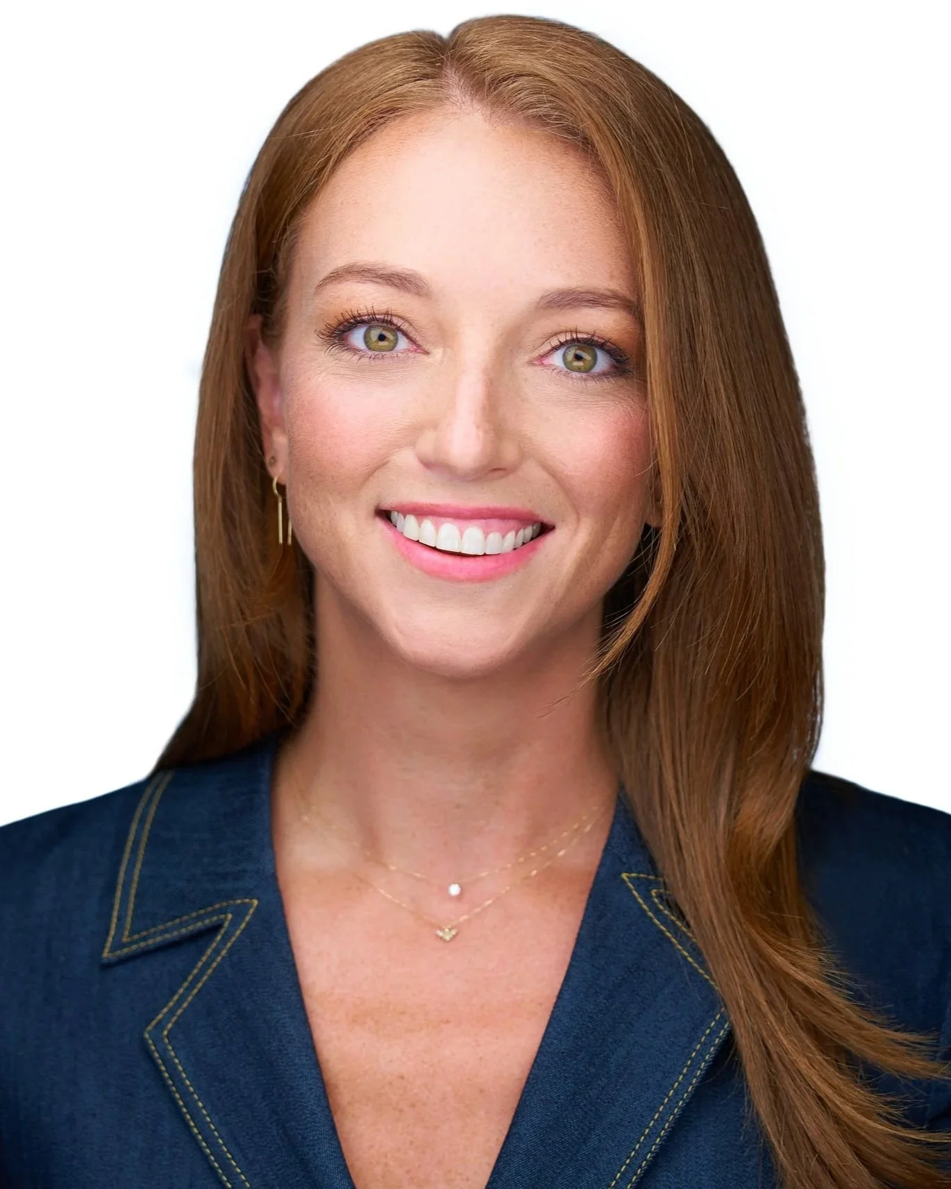 Headshot of a smiling woman with long red hair, green eyes, wearing a navy blue blazer with yellow stitching, a delicate gold necklace, and small gold earrings, against a white background.