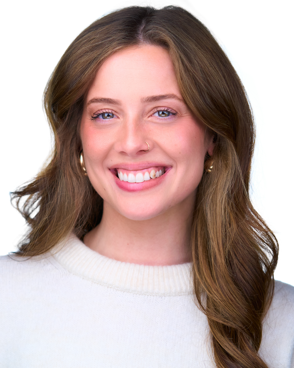 A woman with light brown hair, blue eyes, and a nose piercing, smiling in a white sweater against a plain white background.