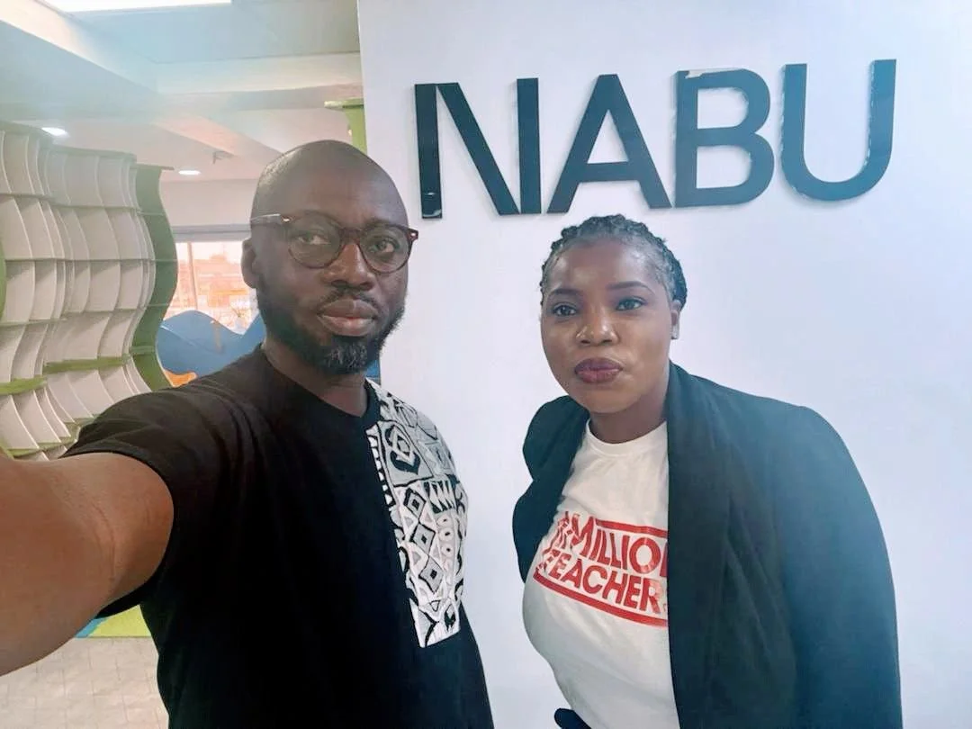 A man and woman taking a selfie together in front of a sign that reads 'NABU'. The man is wearing glasses and a black shirt with white and blue patterns, and the woman is wearing a white t-shirt with red text that says 'Million Teacher' and a black jacket.