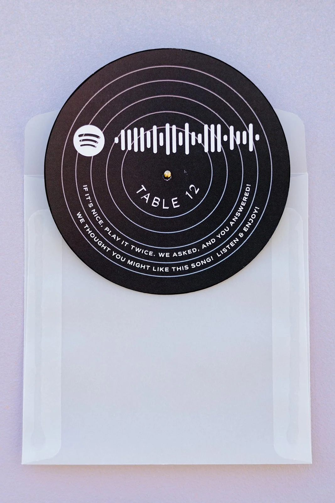 A black circular Spotify-themed paper record label with white text and sound wave pattern, placed on a transparent plastic holder, with the words 'TABLE 12' in the center and a note encouraging guests to play a song they might like.