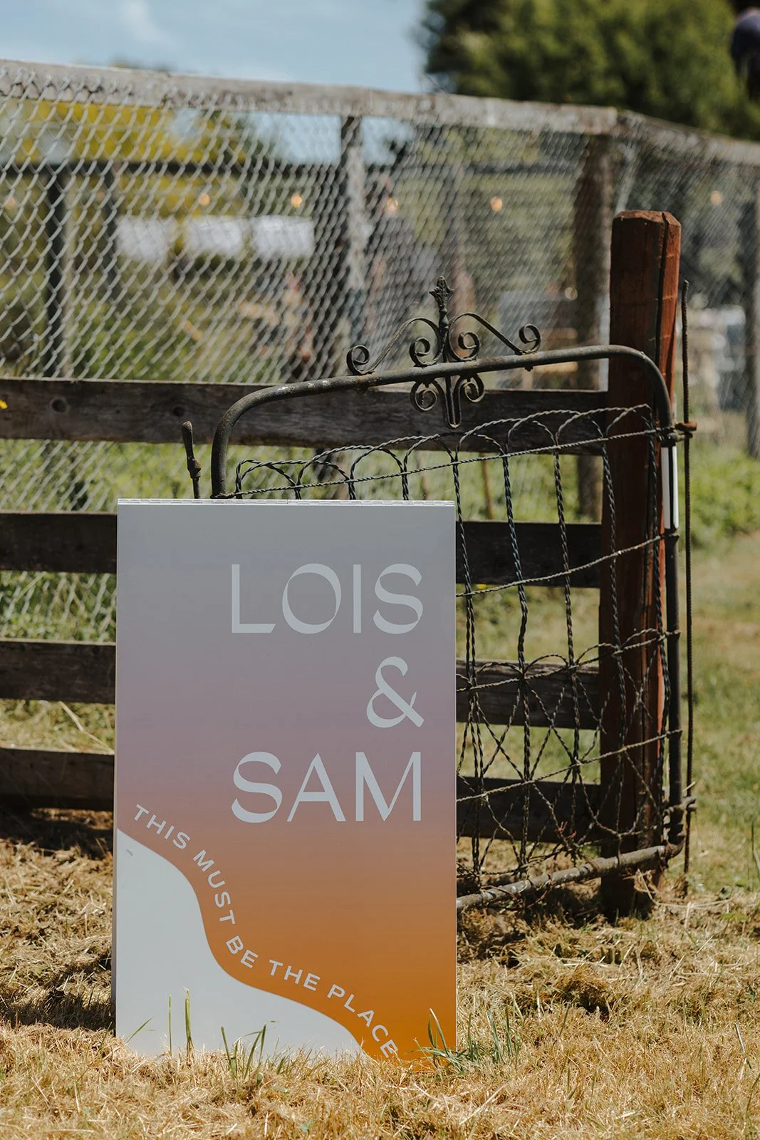 A wedding sign for Lois and Sam with the message "This must be the place," placed outdoors on grass, next to a rustic fence and gate, during daytime with green trees and a blue sky in the background.