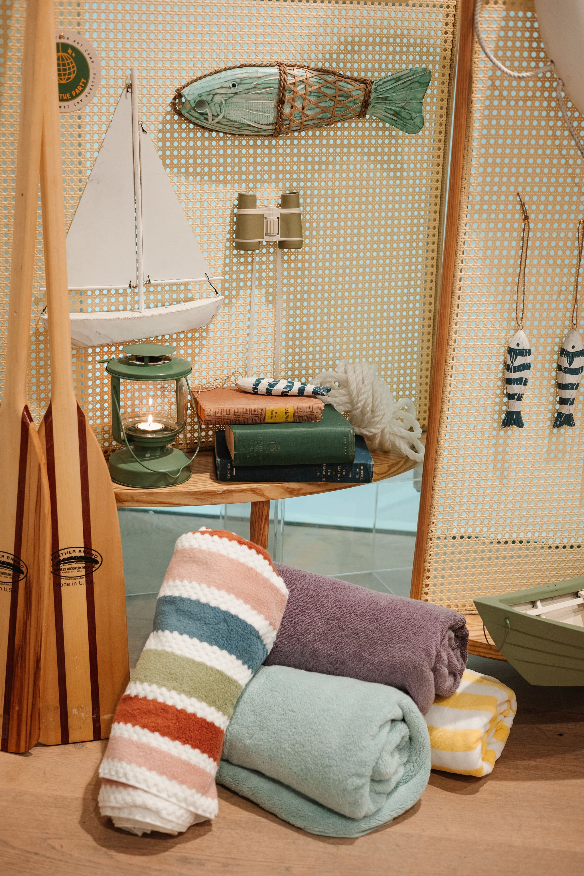 Beach-themed decorative items including wooden oars, rolled towels, a sailboat model, fish decorations, books, a lantern, binoculars, and a fish wall hanging, arranged on and around a wooden table and a screen with a perforated pattern.
