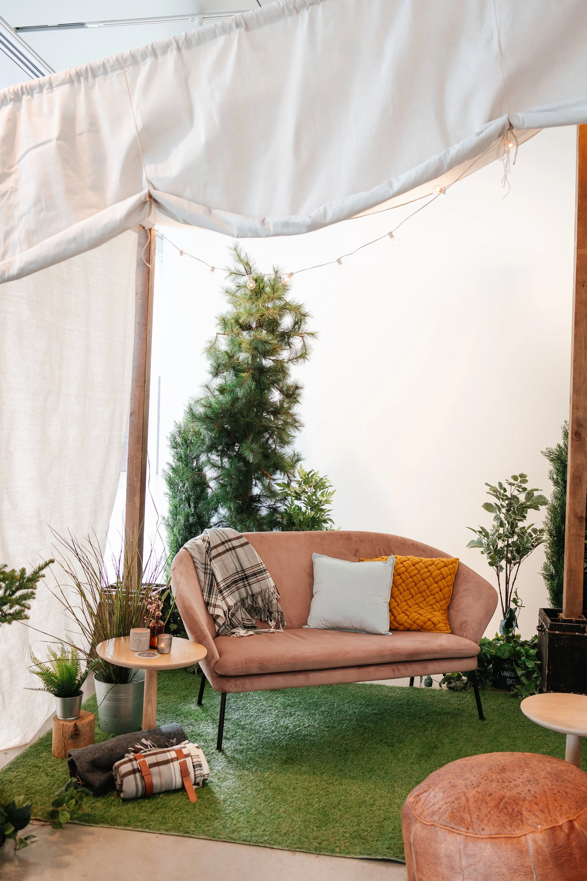 A cozy indoor space with a blush pink sofa, decorative pillows, surrounded by lush green plants, a small wooden side table with candles, and a black-and-white plaid blanket. There is a small artificial grass carpet on the floor, and a tall Christmas