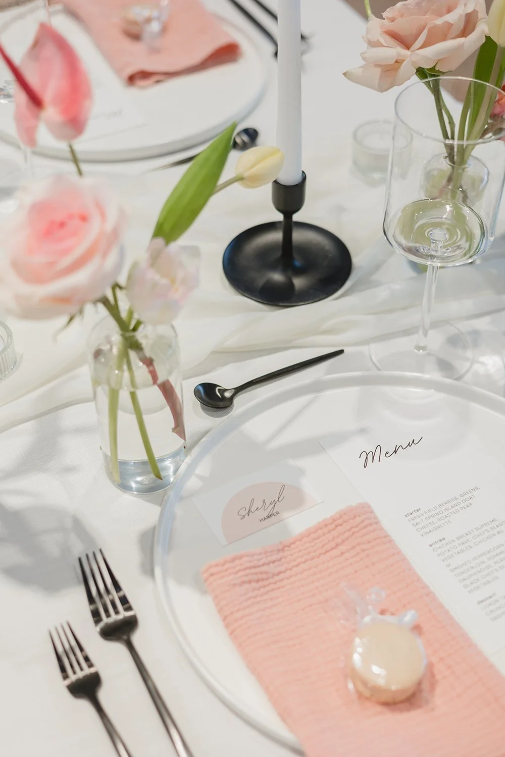 Elegant table setting with pink and white flowers, candles, glassware, silverware, a menu, and a pink napkin with a small round dessert.