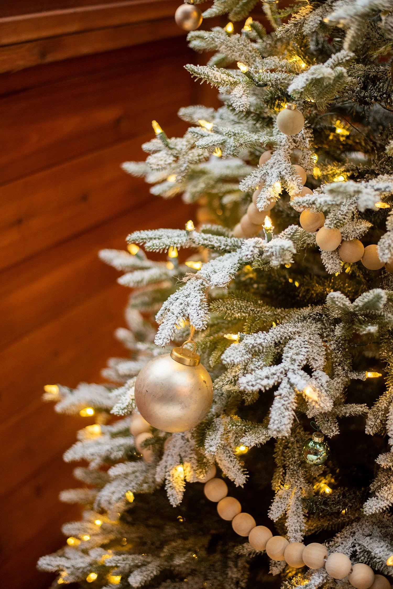 Decorated Christmas tree with snow-flocked branches, gold and white ornaments, warm yellow lights, and a string of wooden beads.