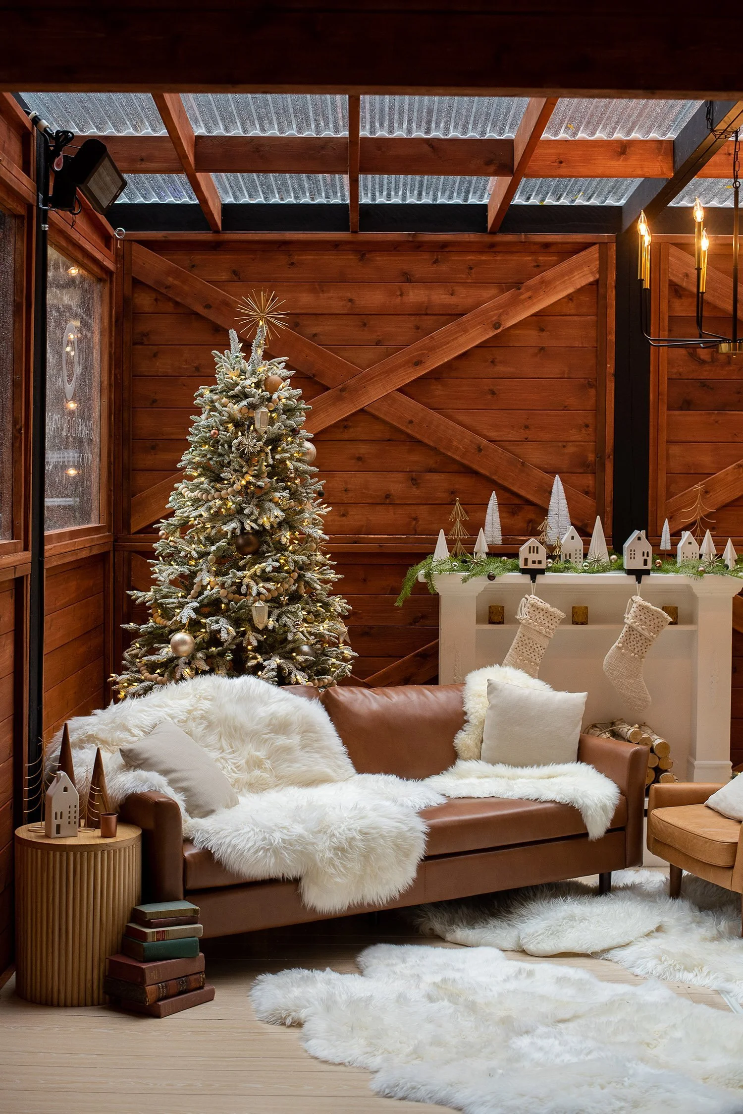 A cozy living room decorated for Christmas with a snow-dusted Christmas tree, stockings hanging on a white mantel, and a plush brown leather sofa covered with white faux fur throws and pillows. There's a small side table with books and decorative items and a fluffy white rug on the wooden floor.
