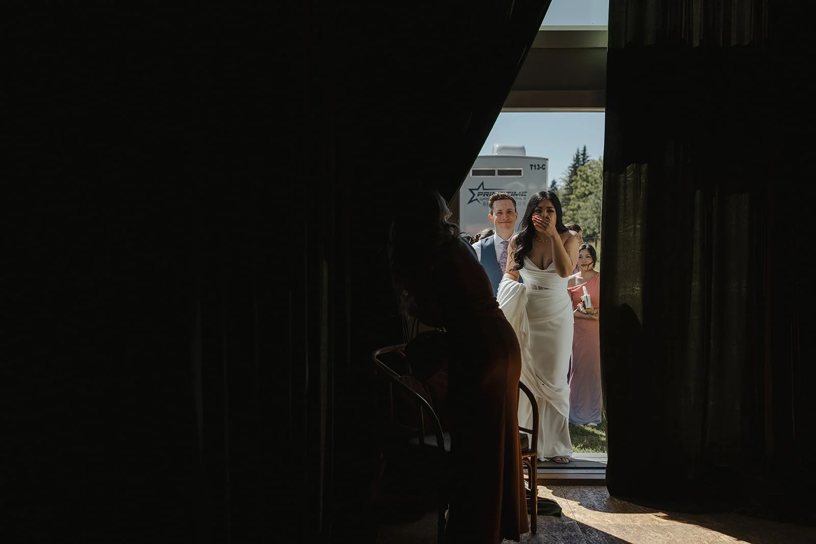 A woman in a white dress is standing near a doorway, covering her mouth with her hand, while a group of people, including a man and two women, are outside. The scene is backlit, creating a silhouette of the woman near the door.