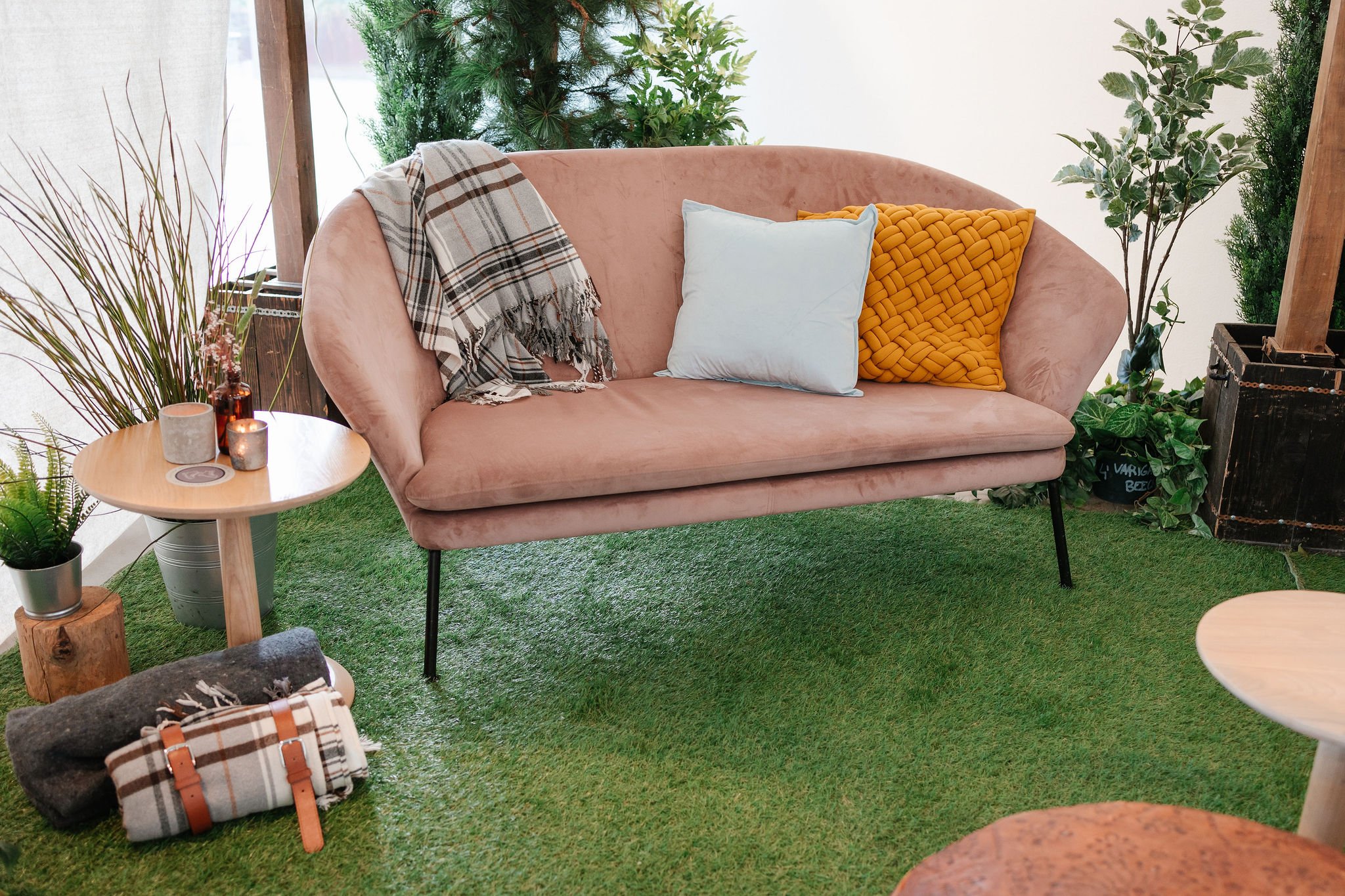 Pink velvet sofa with two decorative pillows, one light blue and one mustard yellow, located in a room with green plants and natural decor. A plaid blanket is draped over the sofa's armrest, and a small round wooden side table with candles and a plan