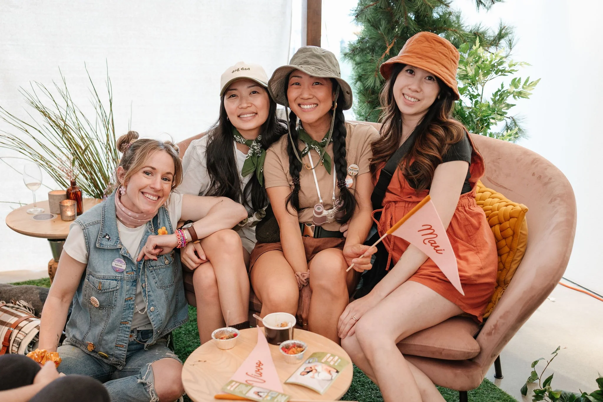 Four smiling women sitting on a pink sofa, one holding a small "Maui" flag, with a small table in front of them holding various snacks. They are outdoors or in a bright, airy space with plants and a Christmas tree in the background.