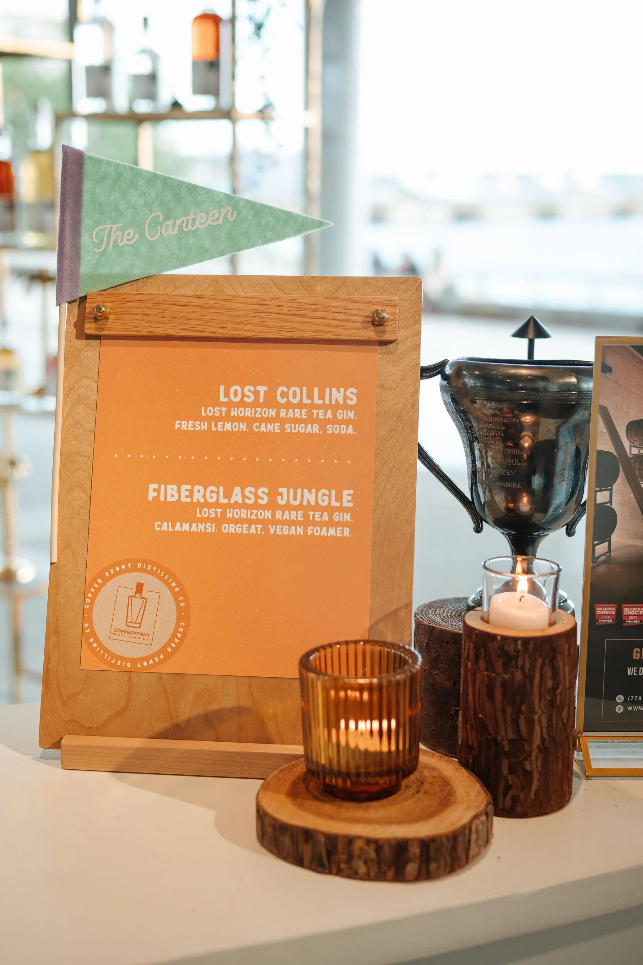 Menu board on a wooden stand with a green flag labeled 'The Canteen', featuring drink options 'Lost Collins' and 'Fiberglass Jungle', with candle holders made of wood and glass, and a black pour-over coffee dripper beside it.