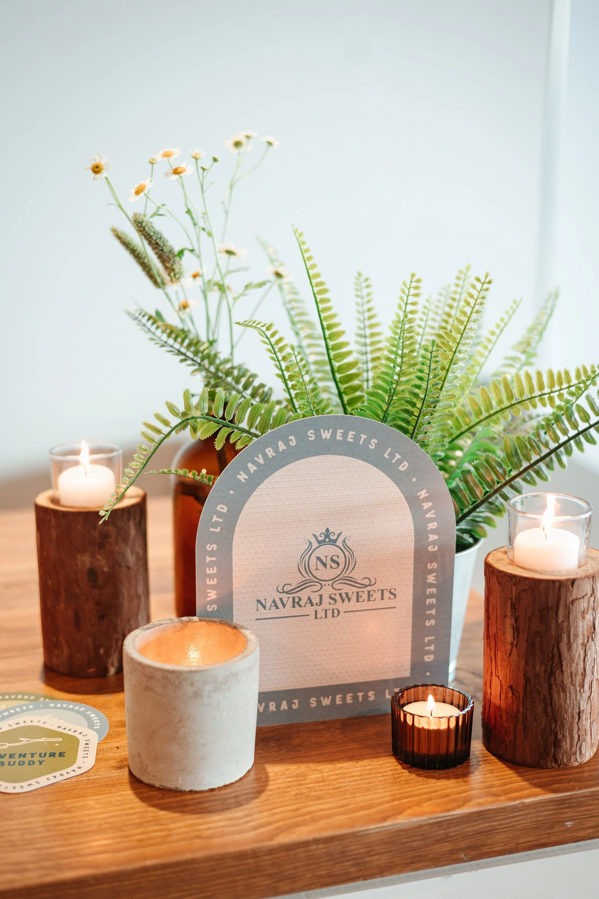 Arrangement of candles in wooden and ceramic holders, a potted fern plant, and a sign that reads 'Navraj Sweets Ltd' on a wooden table, with decorative flowers in the background.