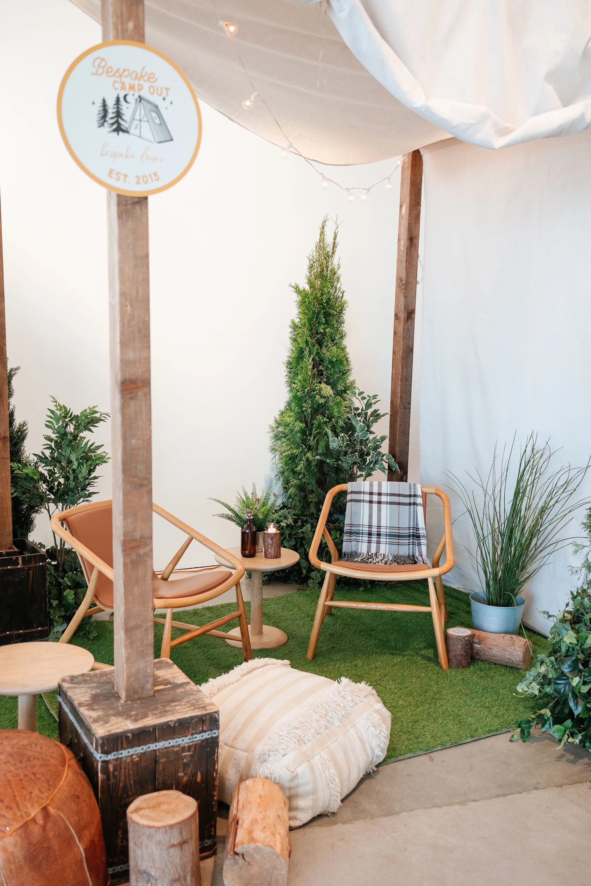 Cozy indoor seating area with two modern wooden chairs, small wooden side tables, potted greenery, a Christmas tree, and a log, with a white sheet backdrop and string lights overhead.