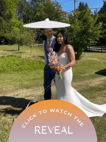 A bride in a white wedding dress holding a bouquet of flowers stands next to a man in a gray suit beneath a white umbrella outdoors with trees and grass in the background.