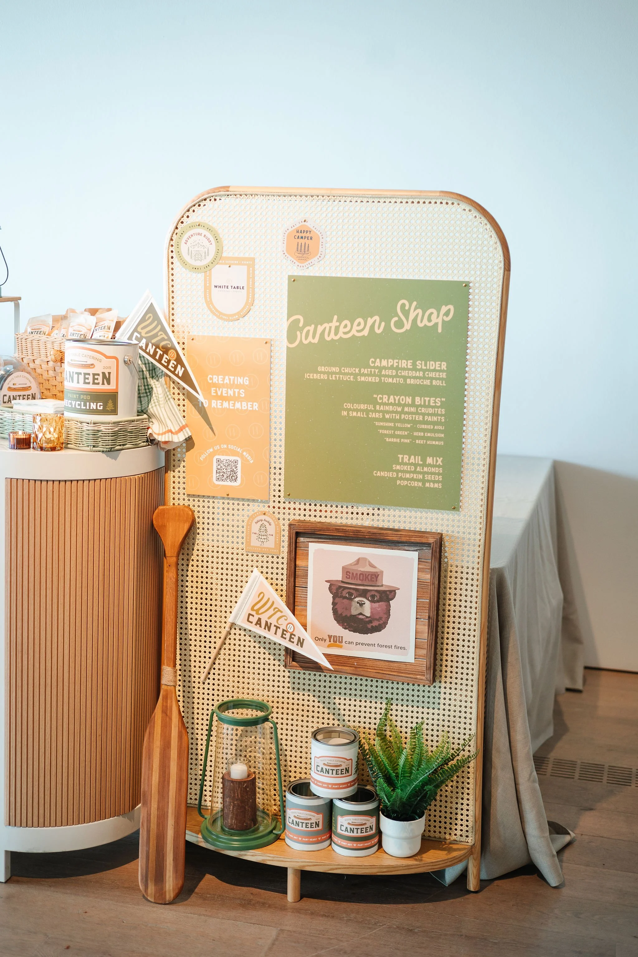 Display board and table with camping-themed items such as cans labeled 'Canteen,' a lantern, a plant, a paddle, and a menu for a camper shop, with signs promoting creating events and forest fire prevention.