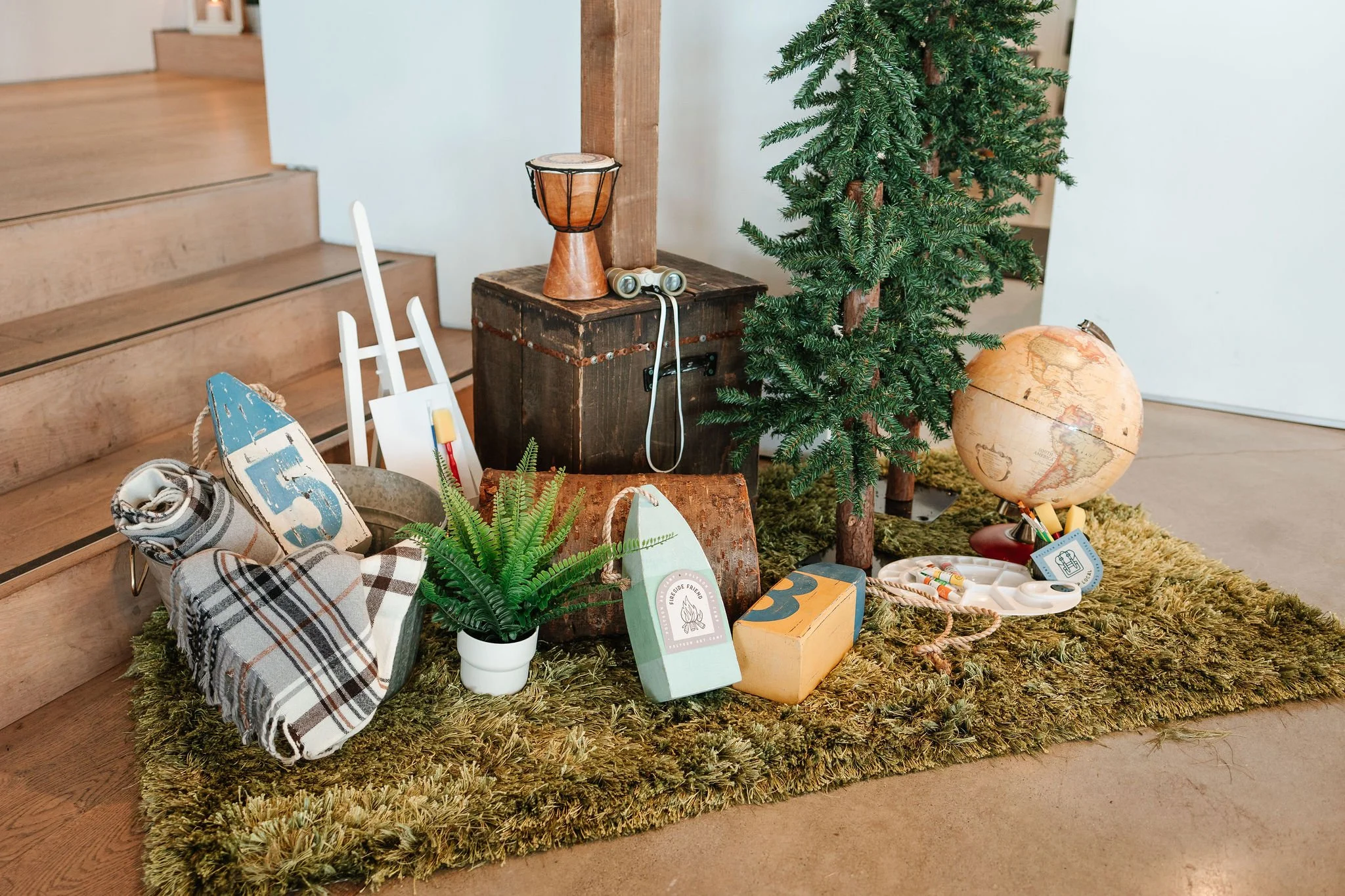 Decorative space with a small artificial Christmas tree, a globe, a vintage suitcase, a wooden box with a drum on top, a potted fern, and vintage-style travel and nautical-themed items on a green shaggy rug near wooden stairs.