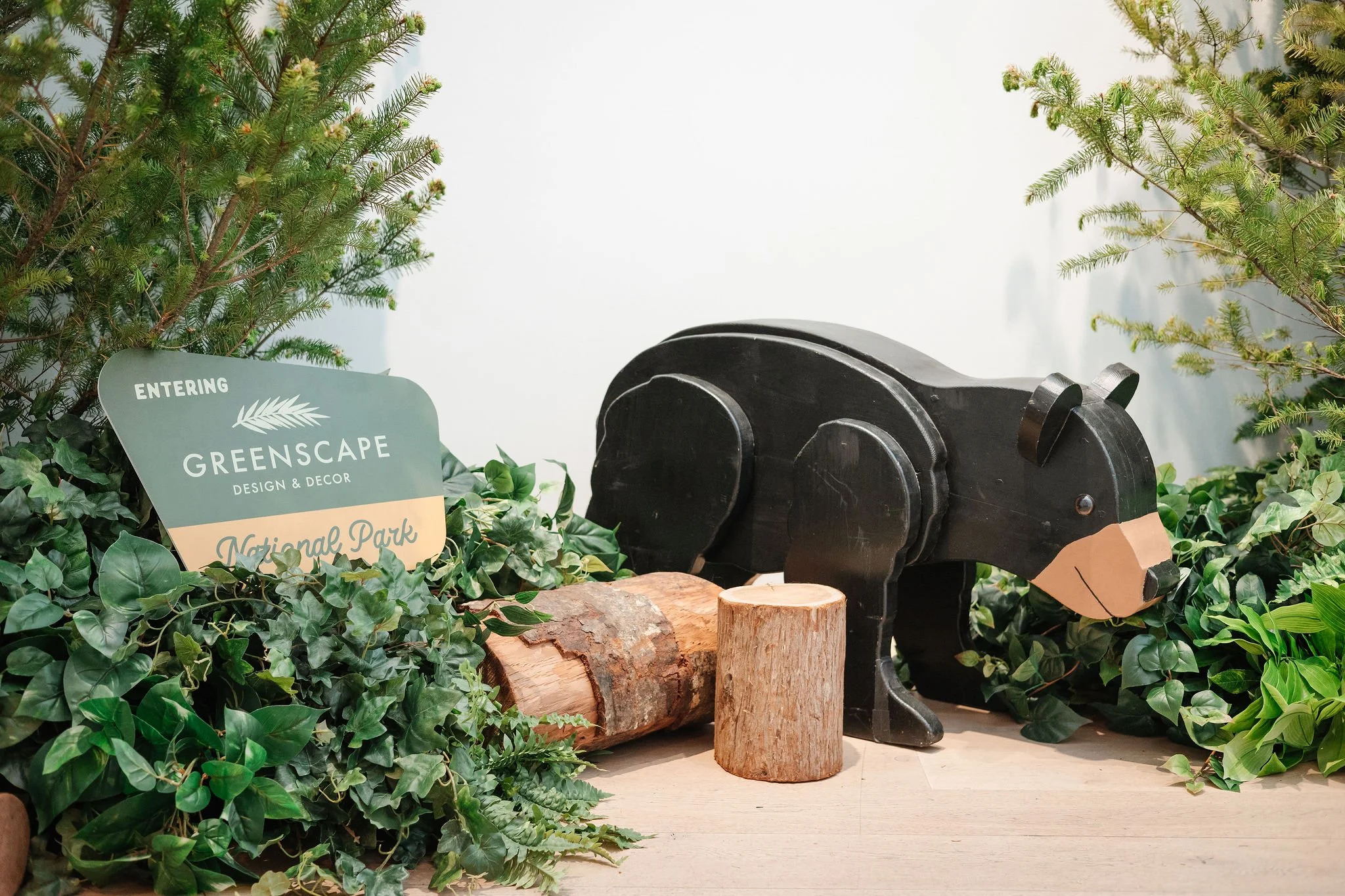 Decorative display with a black wooden bear figure, surrounded by green leafy plants, small tree logs, and a sign that reads 'Entering Greenscape Design & Decor National Park'.