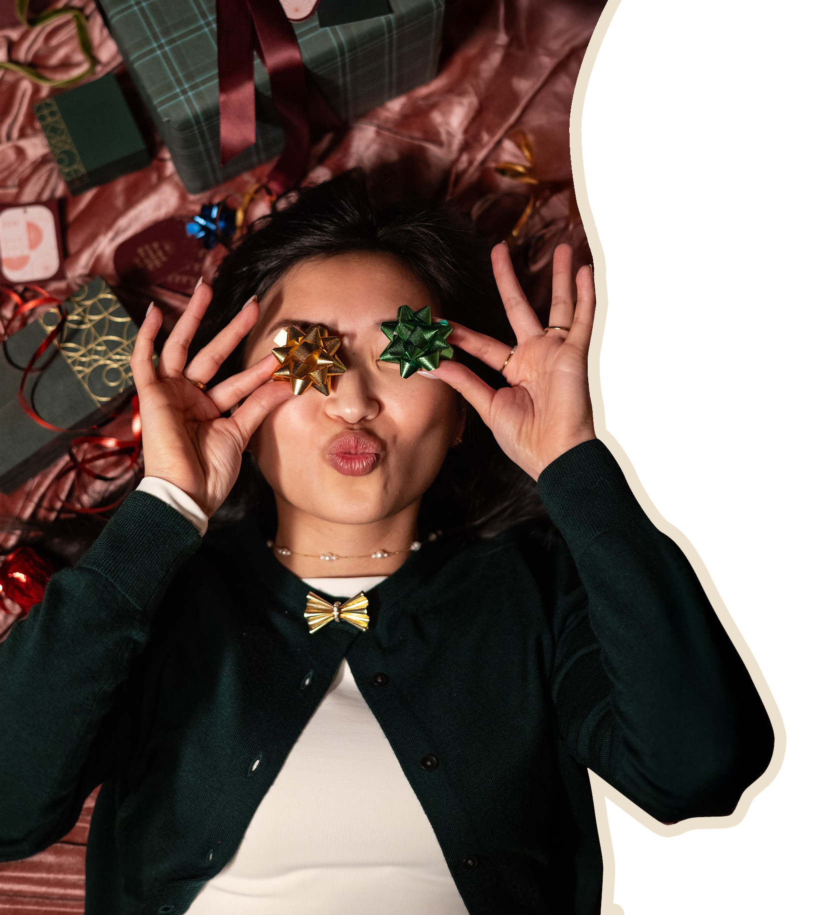 A woman lying on the floor surrounded by wrapped Christmas presents, holding two small gift bows over her eyes, making a playful face.